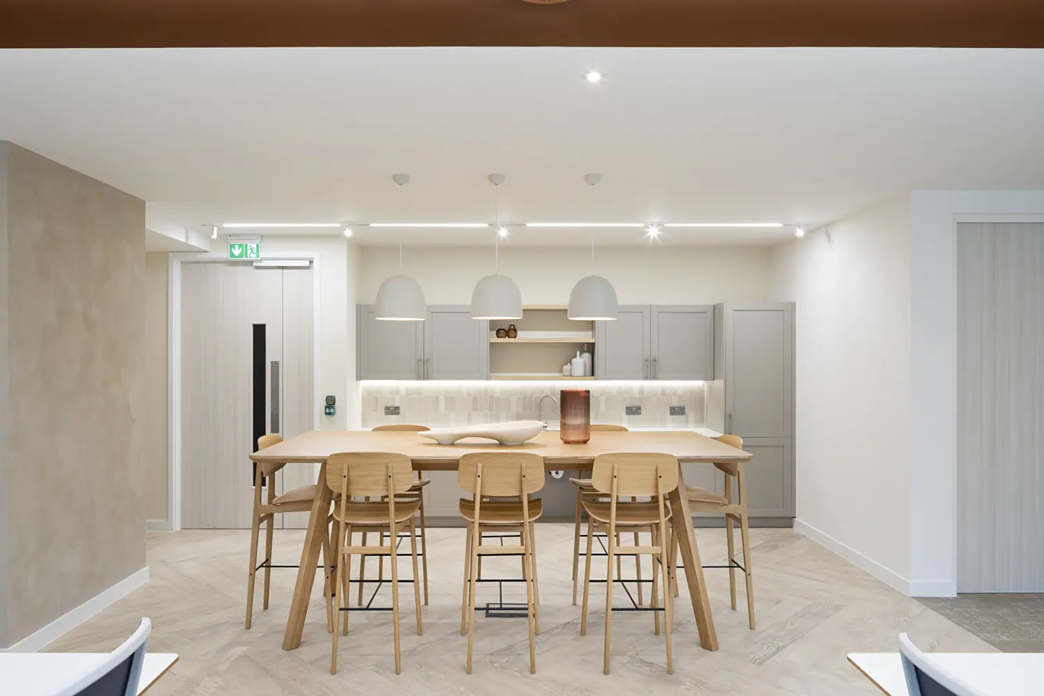 A modern office kitchen and breakout area with a clean, minimalist design. At the centre, a long light wood high table is surrounded by several matching wooden stools with black metal footrests. Above the table, three white dome-shaped pendant lights hang evenly spaced, providing soft, focused lighting.  In the background, a compact kitchenette runs along the wall, featuring light grey cabinetry, open shelving, and a tiled splashback, all illuminated by under-cabinet lighting. To the left, a door with a green emergency exit sign above it is set into a neutral-toned wall. On the right, a white door and wall continue the simple, uncluttered look.  The floor is finished in a light wood-effect pattern, complementing the warm tones of the furniture. Overhead, part of the ceiling reveals exposed services painted in a rich brown colour, framed by a linear LED light. The overall space feels bright, organised and welcoming, with a balanced mix of warm wood, soft neutrals and modern lighting.
