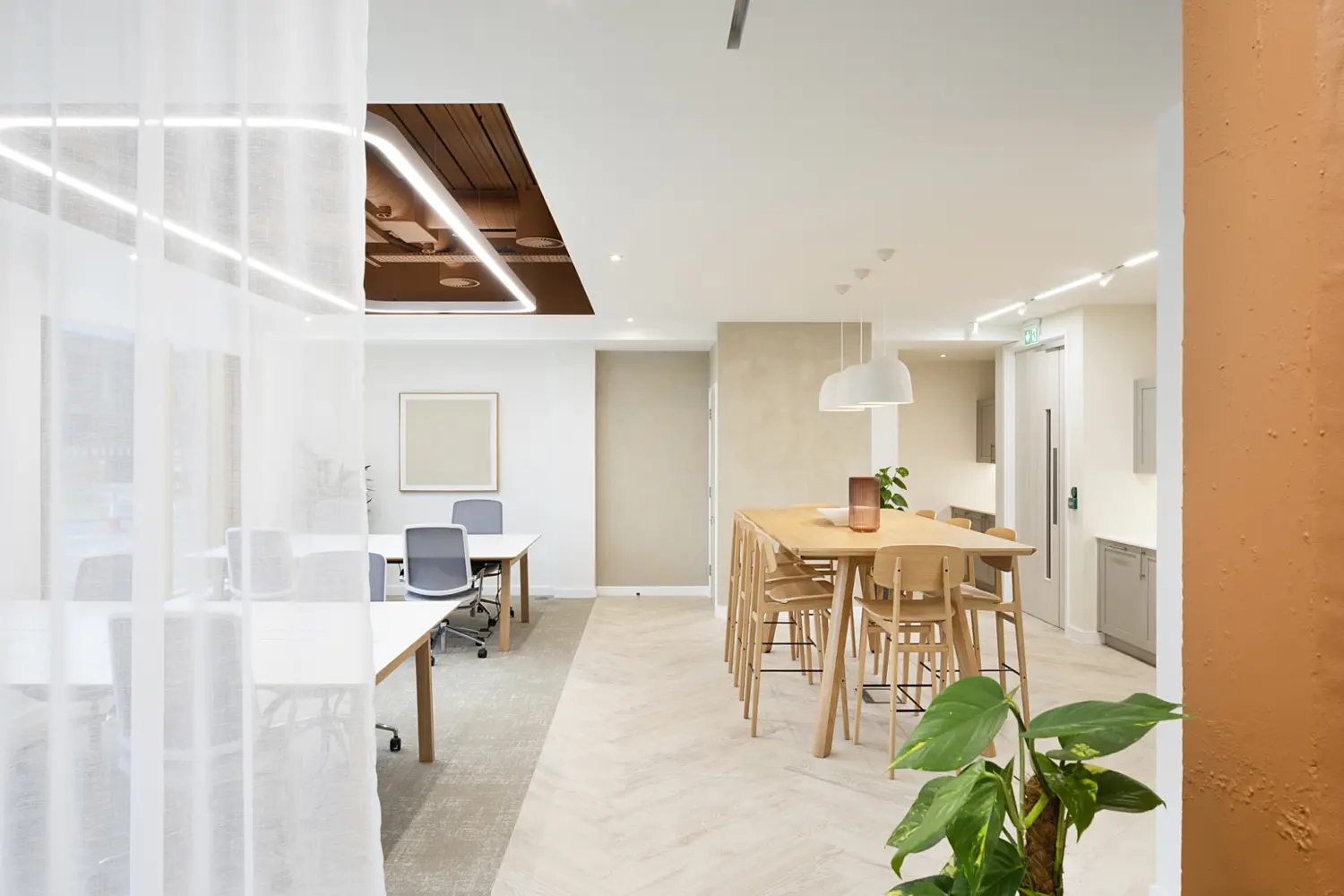 Wide view of a contemporary office interior combining work and breakout spaces. In the foreground on the right, a green potted plant sits beside a textured terracotta-coloured wall. To the left, sheer white floor-to-ceiling curtains partially divide the space, softly diffusing light. Behind the curtains, a work area with white desks and grey office chairs is visible, set on a darker carpeted floor. In the centre-right, a light wood high table with several matching stools forms a casual meeting or social area. Above the table, three white pendant lights hang in a row, while recessed ceiling lights and linear LED strips provide additional illumination. The ceiling includes a feature section with exposed services painted in a warm brown tone, framed by integrated lighting. Further back, neutral-toned walls and cabinetry line the corridor, with a door and small kitchenette-style units visible. The overall space uses a palette of white, beige, warm wood and terracotta accents, complemented by indoor plants and soft lighting to create a bright, modern workplace.
