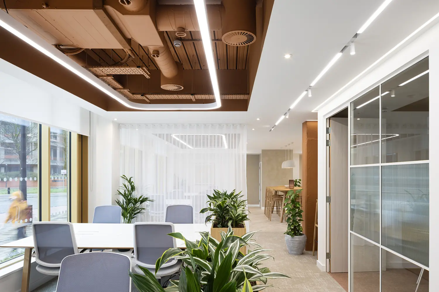 Modern office interior with a clean, minimalist design. In the foreground, a white meeting table is surrounded by several light grey office chairs, with multiple green potted plants placed around the seating area. Large windows on the left let in natural light and show a street scene outside. The ceiling features exposed ductwork painted in a warm brown tone, contrasted with linear LED light strips. In the background, sheer white curtains partially divide the space, leading to a casual breakout area with high wooden stools and a counter. On the right side, a glass-partitioned room with a sliding door is visible. The overall space uses a mix of neutral colours, warm wood accents and indoor greenery to create a bright, contemporary workplace.