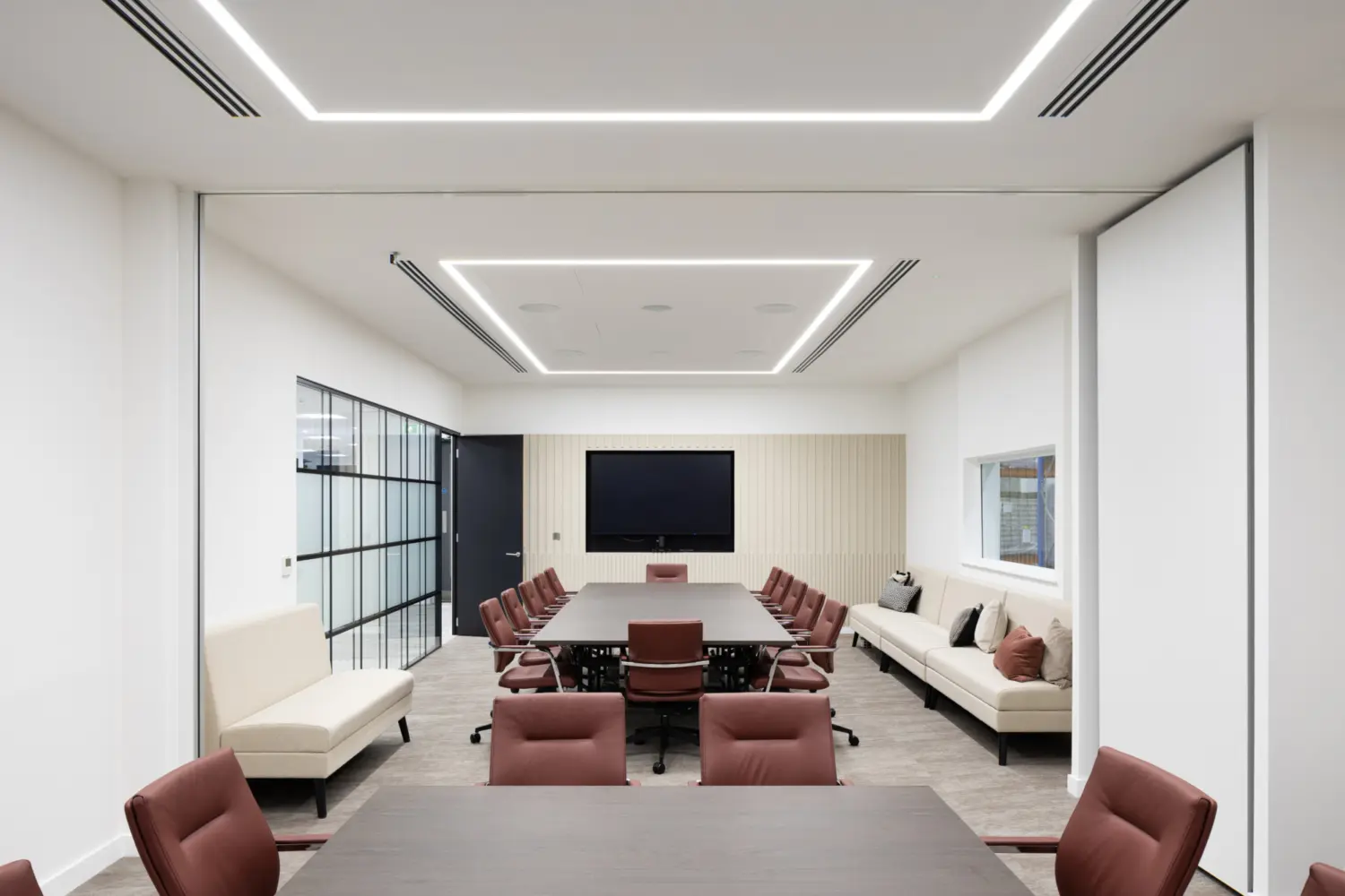 A modern conference room with a large central meeting table surrounded by brown upholstered chairs on castors. At the far end of the room, a wide screen is mounted on a ribbed feature wall. Built‑in sofas with neutral cushions run along both side walls. The room has white walls, a suspended ceiling with integrated rectangular LED lighting, and a glass partition on the left that leads to an adjacent corridor. Sliding partition panels frame the space, and the flooring is a mix of carpet and hard surfaces in muted tones.