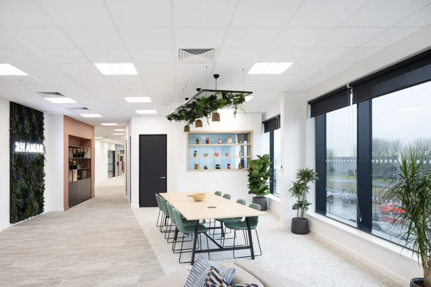 A bright office breakout area with a long light‑wood table and green chairs positioned near large windows with black roller blinds. Above the table hangs a feature light with plants and wicker shades. A shelving unit on the far wall displays assorted jars and containers, with potted plants placed below it. To the left, a corridor leads further into the office and includes a living wall with illuminated signage. The space has white walls, a light carpeted floor and a suspended ceiling with recessed lighting.