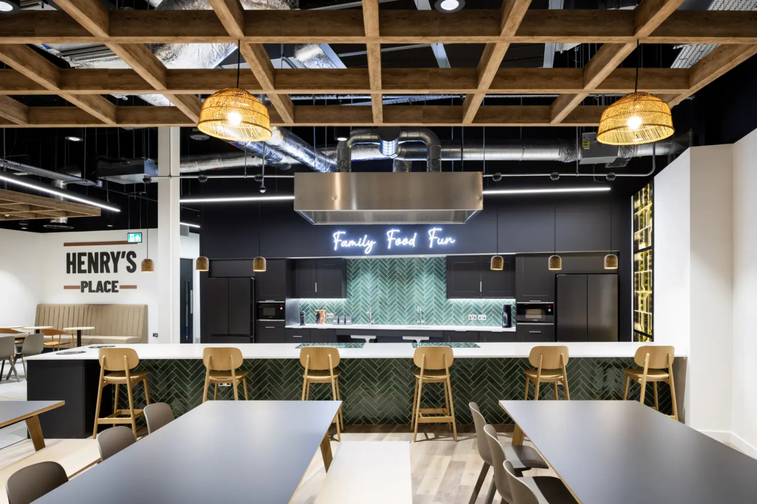 A modern communal kitchen and dining space with long tables and mixed seating in the foreground. At the back is a large kitchen island with wooden bar stools, a green herringbone tiled front and a stainless steel extractor hood above. Behind the island are dark cabinets, built‑in appliances and a green tiled splashback with a neon sign that reads “Family Food Fun”. The ceiling features exposed ductwork and a timber grid structure with woven pendant lights. A sign on the left wall reads “Henry’s Place”.