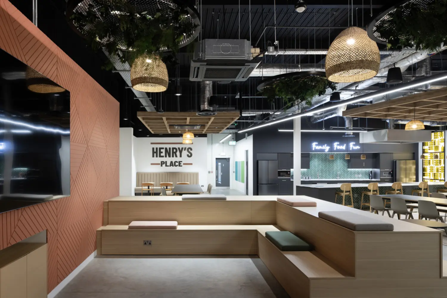 A contemporary breakout space featuring tiered wooden seating with upholstered cushions in muted colours. To the left, a large patterned wall panel frames a mounted screen. The ceiling has exposed ductwork with hanging woven pendant lights and circular planters containing greenery. In the distance, a sign reading “Henry’s Place” is mounted above a small dining area with wooden tables and chairs. Further back, an open kitchen area with a green tiled island and a neon sign reading “Family Food Fun” is visible. The space combines warm timber finishes with industrial ceiling elements and soft seating.Provide your feedback on BizChat
