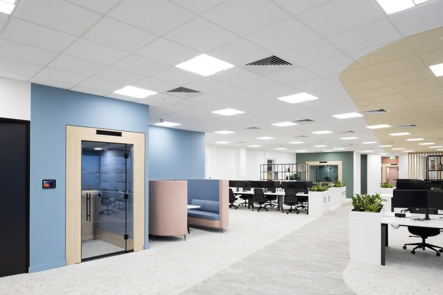 A modern open‑plan office with a light, neutral colour scheme. In the foreground, there is a small enclosed meeting pod with a glass door set within a pale wood frame against a blue wall. Next to it are two high‑backed upholstered booths in soft pastel colours. Beyond this area are clusters of white desks with black office chairs, divided by low planters. The space has a suspended ceiling with recessed lighting, a mix of carpeted and hard flooring and varied wall colours including blue, white and green. The layout is spacious with wide walkways and contemporary finishes throughout.