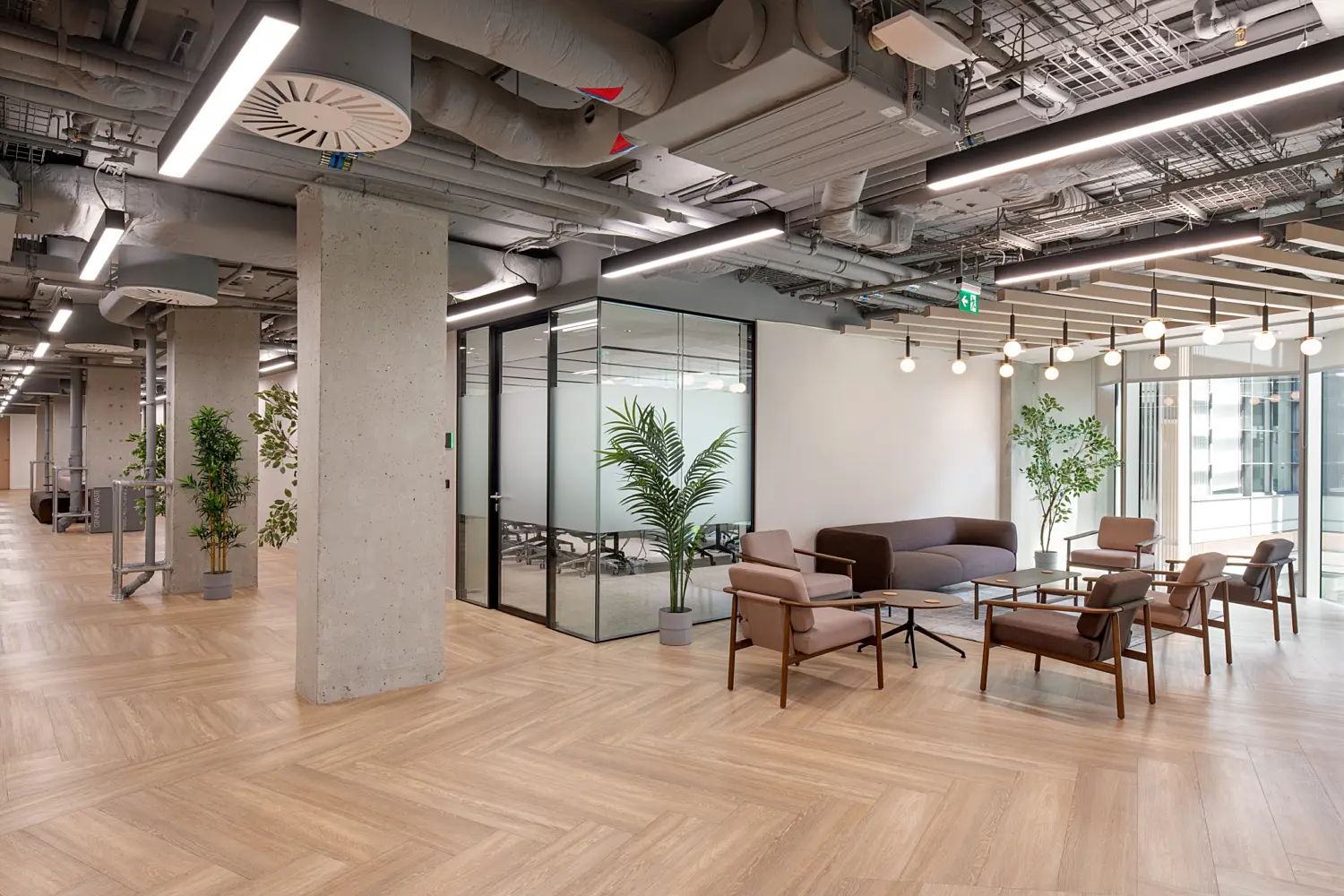 Open-plan office breakout area with lounge seating arranged around low tables beside a glass-fronted meeting room. Large windows bring natural light into the space, while timber-effect flooring, exposed concrete columns and visible ceiling services create a contemporary industrial aesthetic. Pendant lights and integrated linear lighting add warmth, with potted plants placed throughout the workspace to introduce greenery.