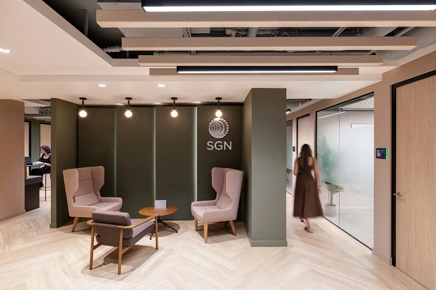 Modern office reception and breakout area featuring lounge seating arranged around a small round table. A textured feature wall displays the SGN logo, while acoustic ceiling panels and pendant lighting create a warm, contemporary atmosphere. Glass-fronted meeting rooms line the corridor, and a person walks through the space towards the offices beyond.