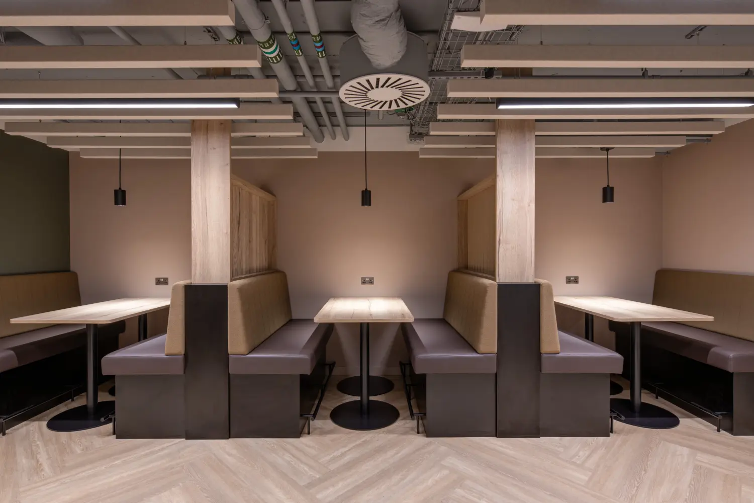 Office breakout space with booth seating arranged around individual tables for informal meetings and focused work. Upholstered benches, timber finishes and soft neutral colours create a comfortable setting, while suspended acoustic ceiling panels and integrated lighting contribute to a calm, contemporary environment. Exposed ceiling services and herringbone flooring add a modern workplace aesthetic.