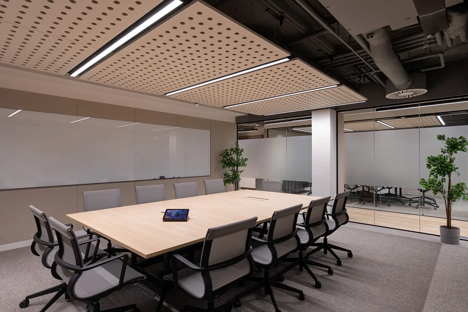 Modern meeting room featuring a large boardroom table surrounded by ergonomic task chairs. A full-width whiteboard spans one wall, while glazed partitions provide views into adjacent meeting spaces. Acoustic ceiling panels and suspended lighting create a comfortable environment for collaboration, with potted plants adding greenery to the contemporary workspace.