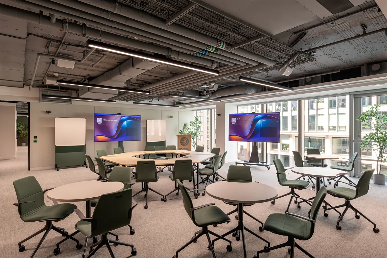 Flexible training and collaboration space with movable tables and green task chairs arranged for group work. Two large presentation screens display branded content, while mobile whiteboards provide additional space for workshops and meetings. Floor-to-ceiling windows bring natural light into the room, and exposed ceiling services with contemporary lighting create a modern workplace environment designed for learning and collaboration.