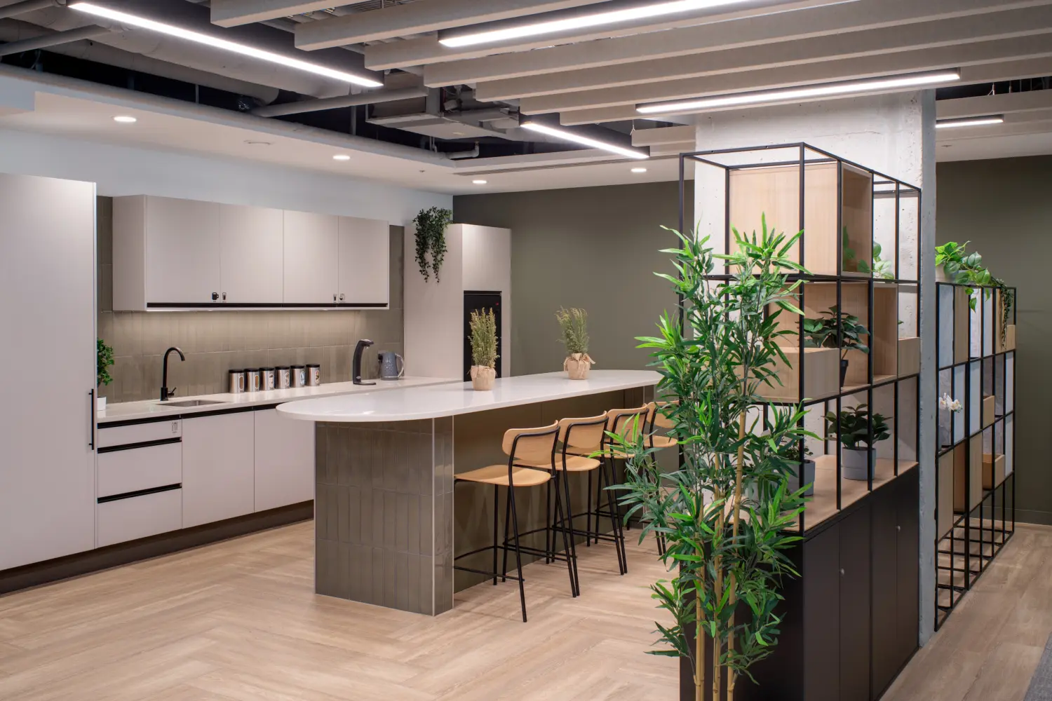 Contemporary office tea point featuring a central island with bar stools, integrated appliances and modern cabinetry. Open shelving units filled with plants create a natural divider within the space, while timber finishes, greenery and neutral tones contribute to a warm, welcoming atmosphere. Linear lighting and acoustic ceiling features complement the modern workplace design.