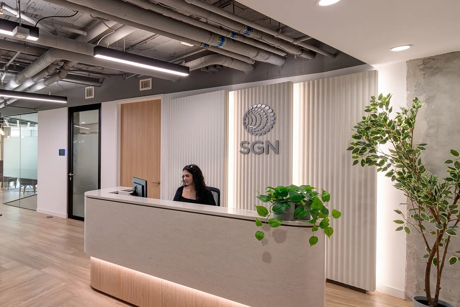 Modern office reception area featuring a curved stone-fronted desk with integrated lighting and timber detailing. A textured feature wall behind the desk displays the SGN logo, while planting and natural wood flooring add warmth to the space. Glass-fronted meeting rooms sit adjacent to the reception area, and exposed ceiling services with contemporary lighting create a modern workplace aesthetic.
