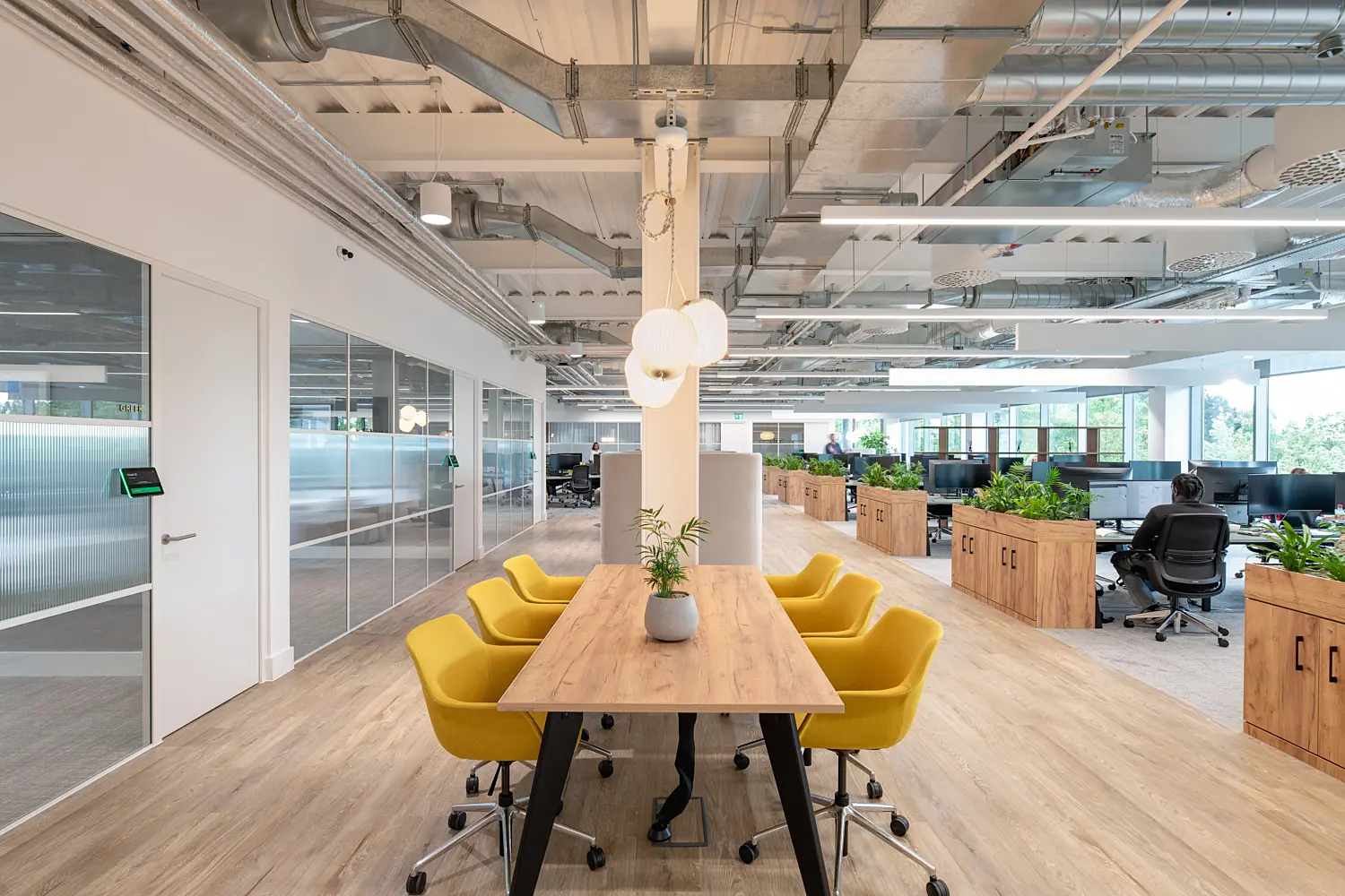 Long, light-filled open-plan office featuring a central wooden meeting table surrounded by six upholstered yellow swivel chairs on wheels. Above the table hang three spherical pendant lights attached to a white structural column. The ceiling is exposed, showing silver ductwork, pipes and suspended linear lighting.  To the left, a row of glass-partitioned meeting rooms with sliding doors runs along the wall, with frosted lower panels and clear upper sections. To the right and extending into the background, rows of desks with computer monitors and office chairs are divided by rectangular wooden planters filled with green plants.  The floor combines light wood in the central walkway with carpeted areas around the workstations. Large windows at the far end allow natural light into the space, and additional potted plants add greenery throughout. The overall setting is modern, clean and airy with a mix of neutral tones and bright accents.