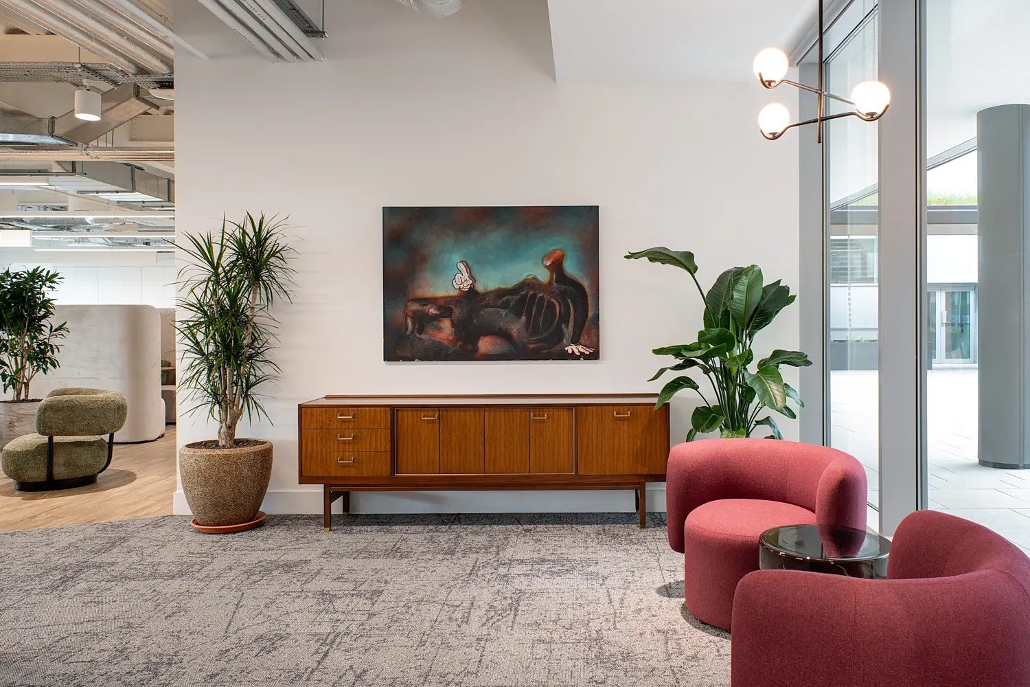 Contemporary breakout seating area within an open-plan office, combining soft furnishings, natural materials and artwork. In the foreground to the right, two rounded upholstered armchairs in a muted red tone are positioned around a small circular side table with a dark top. A large floor-to-ceiling window runs along the right-hand side, bringing in natural light and views of an external courtyard or neighbouring building.  At the centre of the scene, a mid-century style wooden sideboard with multiple drawers and cabinet doors sits against a white wall. Above the sideboard, a large abstract painting in deep, moody tones adds contrast and visual focus. Tall potted plants with broad and slender leaves are placed on either side of the sideboard, introducing greenery into the space.  Overhead, a wall-mounted light fixture with multiple spherical bulbs extends from the right wall, complementing the soft curves of the seating. The floor is finished with a textured grey carpet, while the background to the left reveals part of the wider office, including exposed ceiling services, additional seating and partitioning. The overall setting is calm, modern and designed for informal meetings or relaxation within the workplace.