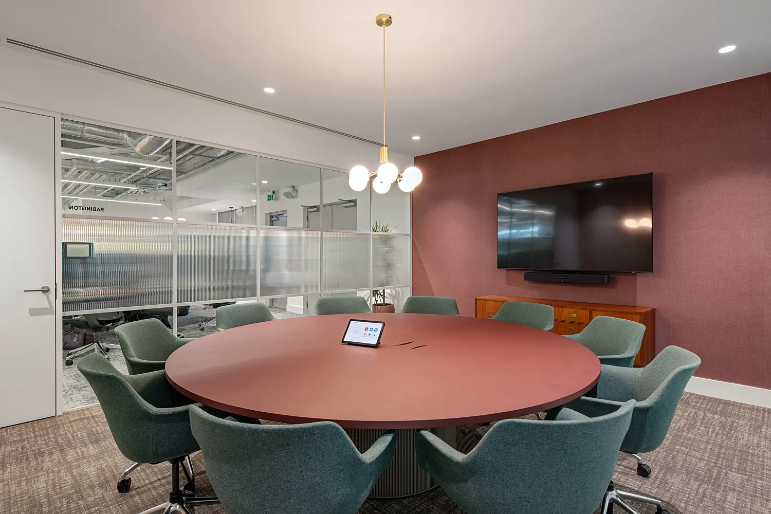 Contemporary meeting room with a round, muted red table at the centre, surrounded by eight upholstered swivel chairs in a soft green tone on castors. A small touchscreen meeting control panel sits in the middle of the table. Above, a pendant light with multiple spherical bulbs hangs from the ceiling, providing warm, even illumination.  The right-hand wall is finished in a textured, deep red surface and features a large wall-mounted flat screen with a soundbar below, positioned above a low wooden sideboard. To the left, a glass partition wall with frosted lower panels and clear upper sections separates the room from the open office beyond, where desks, chairs and exposed ceiling services are visible.  The space is finished with neutral carpeting, white ceilings with recessed lighting and clean-lined detailing throughout. A small potted plant sits near the glass wall, adding greenery to the otherwise structured, modern setting.