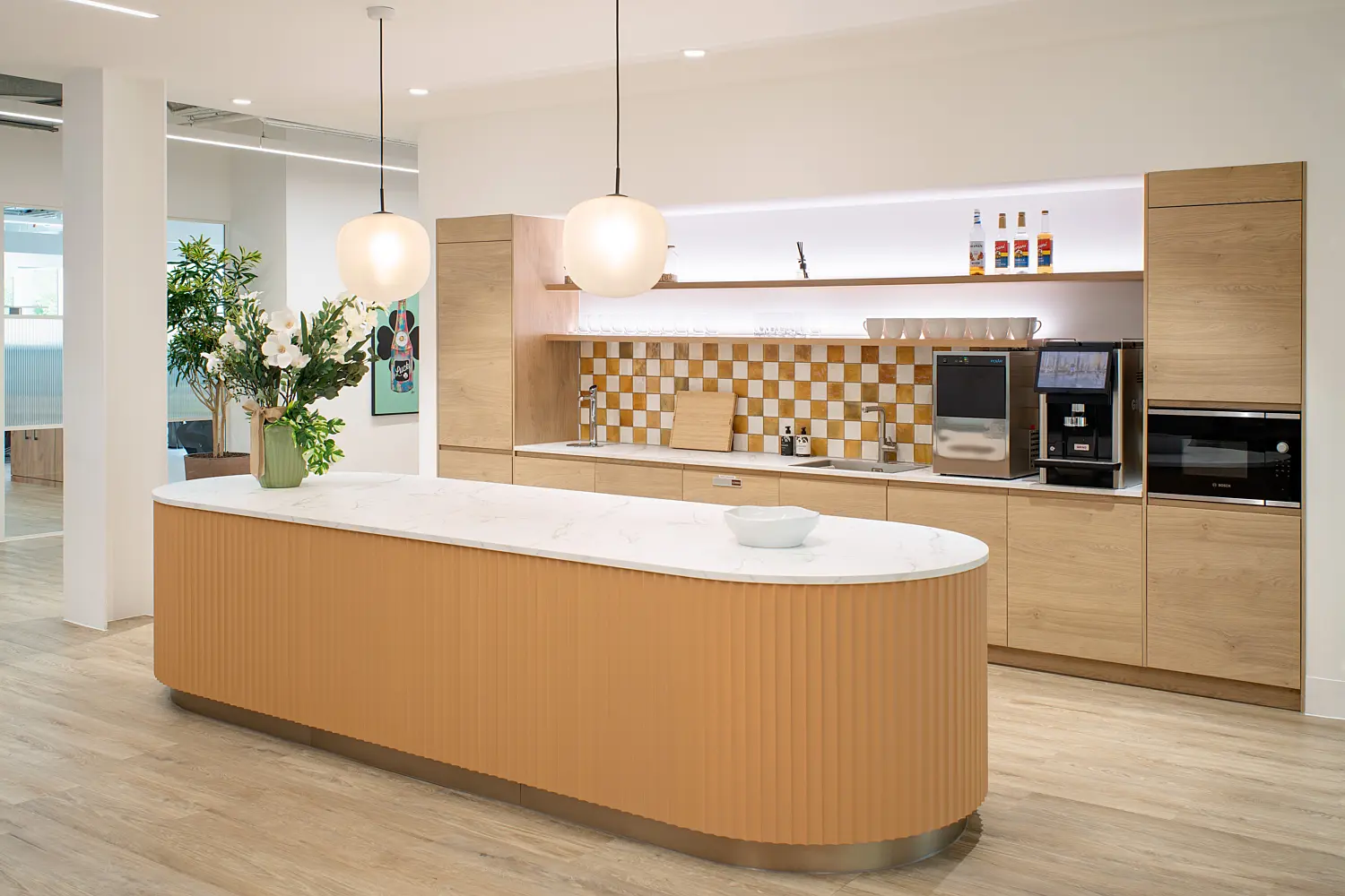 Modern office kitchenette and breakout space with a clean, minimalist design. In the foreground, a large curved island counter with a fluted wood front and a white stone worktop sits on light wood flooring, with a small bowl and a tall vase of flowers placed on top. Two round pendant lights hang above the island, casting soft, even light.  Behind the island, a wall of light wood cabinetry integrates appliances including a built-in oven and a coffee machine. Open shelves display glassware, mugs and bottles, while a warm-toned, checkered tile backsplash adds visual interest. A sink and preparation area run along the counter beneath the shelving.  To the left, the space opens towards the wider office, with a glimpse of glass partitions, additional greenery and wall art. The overall palette combines light timber, white surfaces and warm accents, creating an inviting, contemporary feel.
