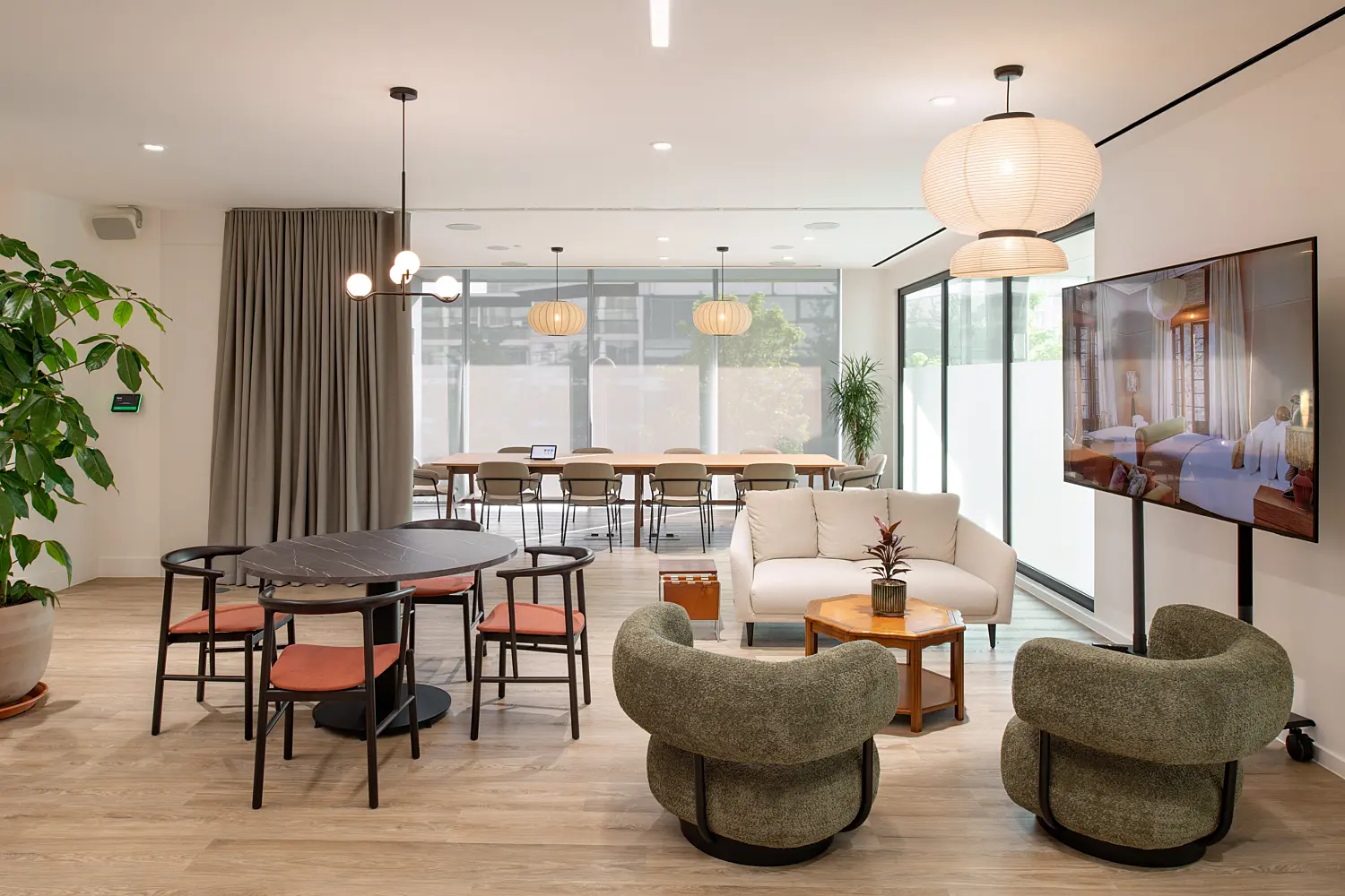 Modern breakout and collaboration area within an office, combining lounge seating and shared workspaces. In the foreground, two rounded upholstered armchairs in a muted green tone face a small wooden coffee table with a plant on top, alongside a light-coloured sofa and an adjacent side table. To the left, a circular dark table is surrounded by four chairs with wooden frames and muted red seat pads, creating a more casual meeting spot.  To the right, a large freestanding digital display screen sits on a stand, positioned near the seating area. Above, a mix of pendant lights hangs from the ceiling, including sculptural multi-globe fixtures and larger paper-style lantern shades, adding soft, warm lighting.  In the background, a long communal table with multiple chairs sits in front of full-height windows with frosted lower sections, allowing natural light to fill the space while maintaining privacy. Floor-to-ceiling curtains line the left side, and a tall potted plant adds greenery near the edge of the frame.  The space features light wood flooring, white walls and a clean, contemporary finish, blending informal seating with functional work areas for meetings or relaxation.