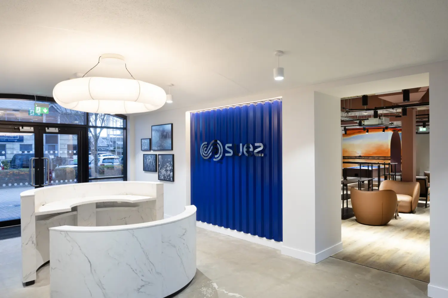 Reception area with a curved white marble reception desk set on polished concrete flooring beneath sculptural pendant lighting. A blue feature wall with vertical panelling displays the SUEZ logo in white, with framed artwork mounted on the adjacent wall. Glazed entrance doors sit to the left, while an opening on the right reveals a breakout space with soft seating, timber flooring and a mural featuring arched forms and a sunset scene.