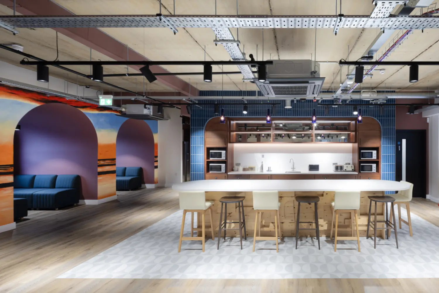 Modern communal kitchen and seating area with a large white island and wooden stools at the centre, exposed ceiling services above and timber flooring throughout. Arched wall recesses with bench seating and a sunset mural line the left side, while built in appliances and blue tiled walls frame the kitchen at the back.