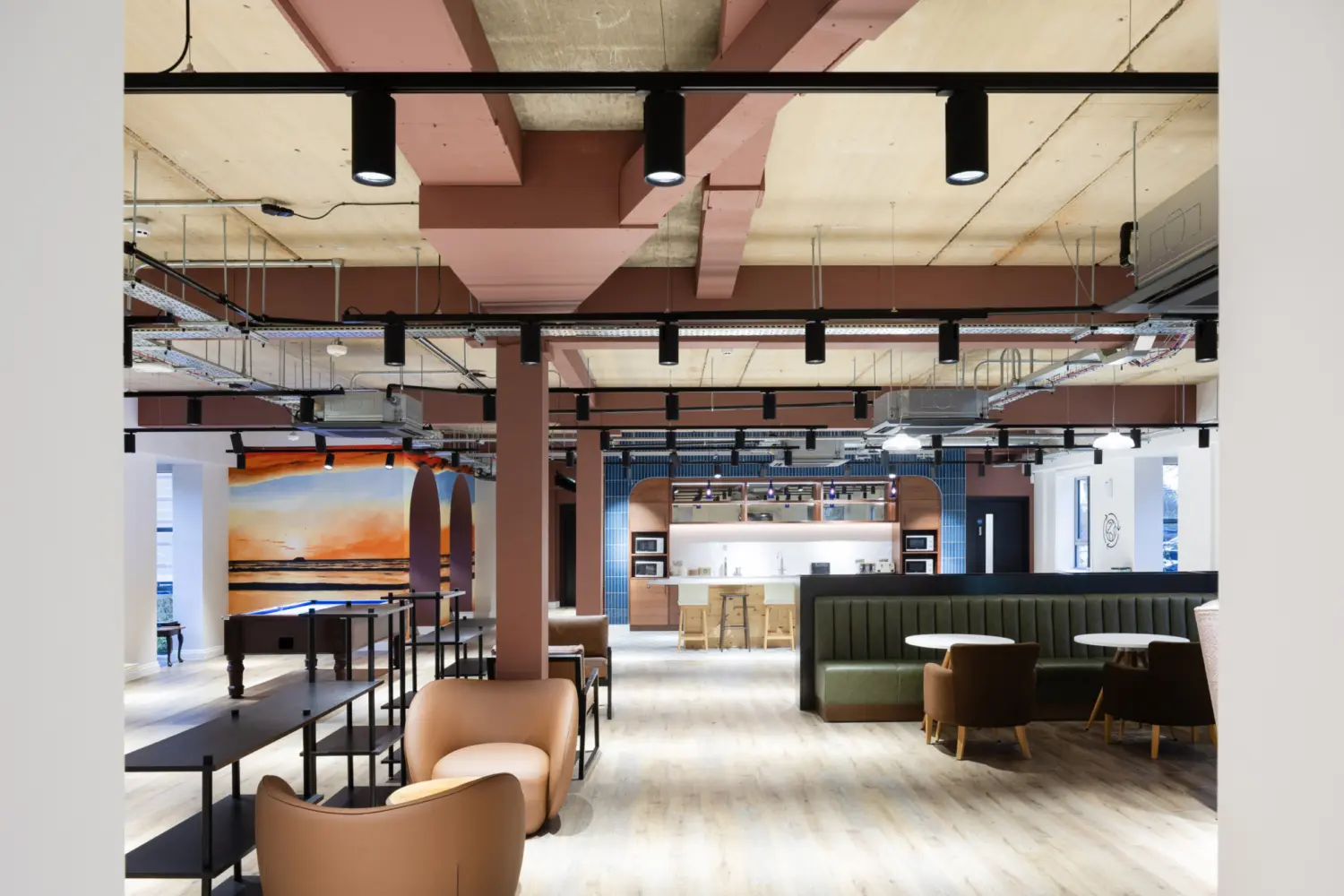 Wide view of an open plan breakout space with timber flooring, exposed ceiling services and suspended lighting. Soft seating and small tables sit in the foreground, leading towards a communal kitchen with a white island and stools at the back. A pool table and wall mural with arched forms and a sunset scene appear on the left, while booth seating and armchairs define informal meeting areas on the right.