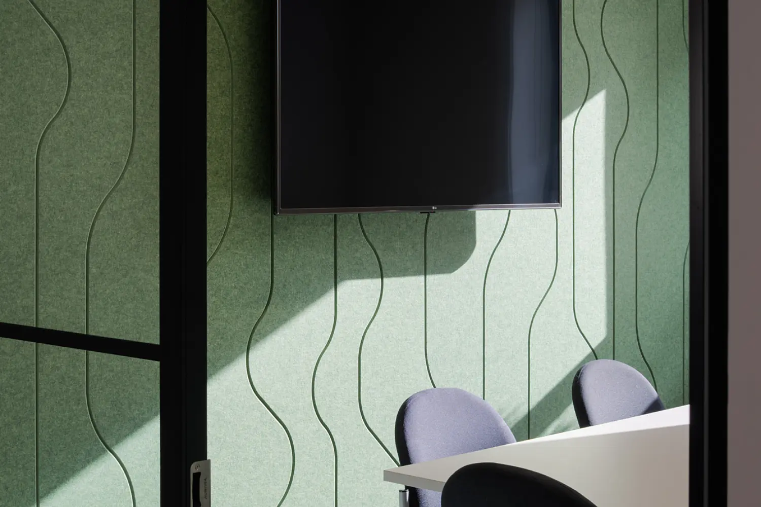 Small meeting room viewed through a glass door, with green acoustic wall panels patterned with vertical waves, a wall-mounted black screen, a white table and four upholstered meeting chairs. Overhead curved ceiling panels and natural light cast shadows across the walls and green carpet.