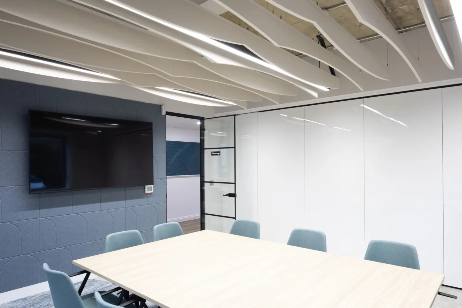 Contemporary meeting room with a large light wood table surrounded by upholstered chairs in muted blue tones. A wall mounted screen sits on a textured blue feature wall to the left, while full height white writable panels line the right side. Overhead, sculptural acoustic baffles with integrated linear lighting are suspended beneath an exposed ceiling, creating a clean, modern workspace.