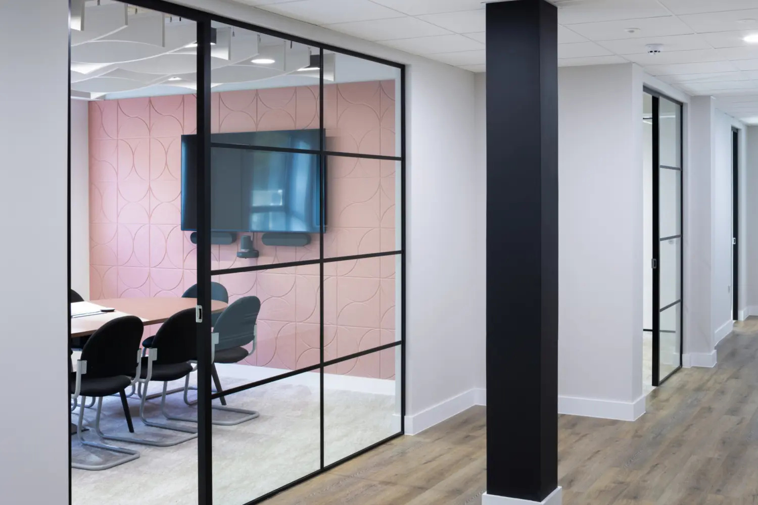 Office corridor with timber effect flooring and white walls, featuring a glass fronted meeting room on the left with black metal framing. Inside the meeting room, a wall mounted screen, table and upholstered chairs sit against a soft pink patterned feature wall. Recessed ceiling lighting runs through the corridor, with additional meeting rooms visible further along the hallway.