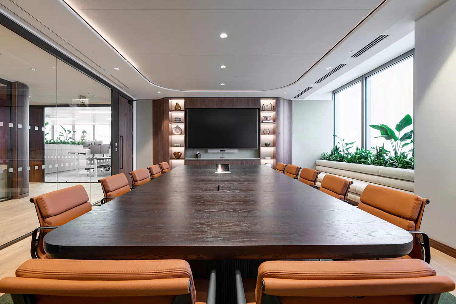 A modern conference room featuring a large wooden table surrounded by brown leather chairs. One side of the room is enclosed with glass walls, while the far end features a wall-mounted flat-screen television and built-in shelving displaying decorative items. Large windows along one side let in natural light and are adorned with potted plants on the sills, adding a touch of greenery to the professional setting.
