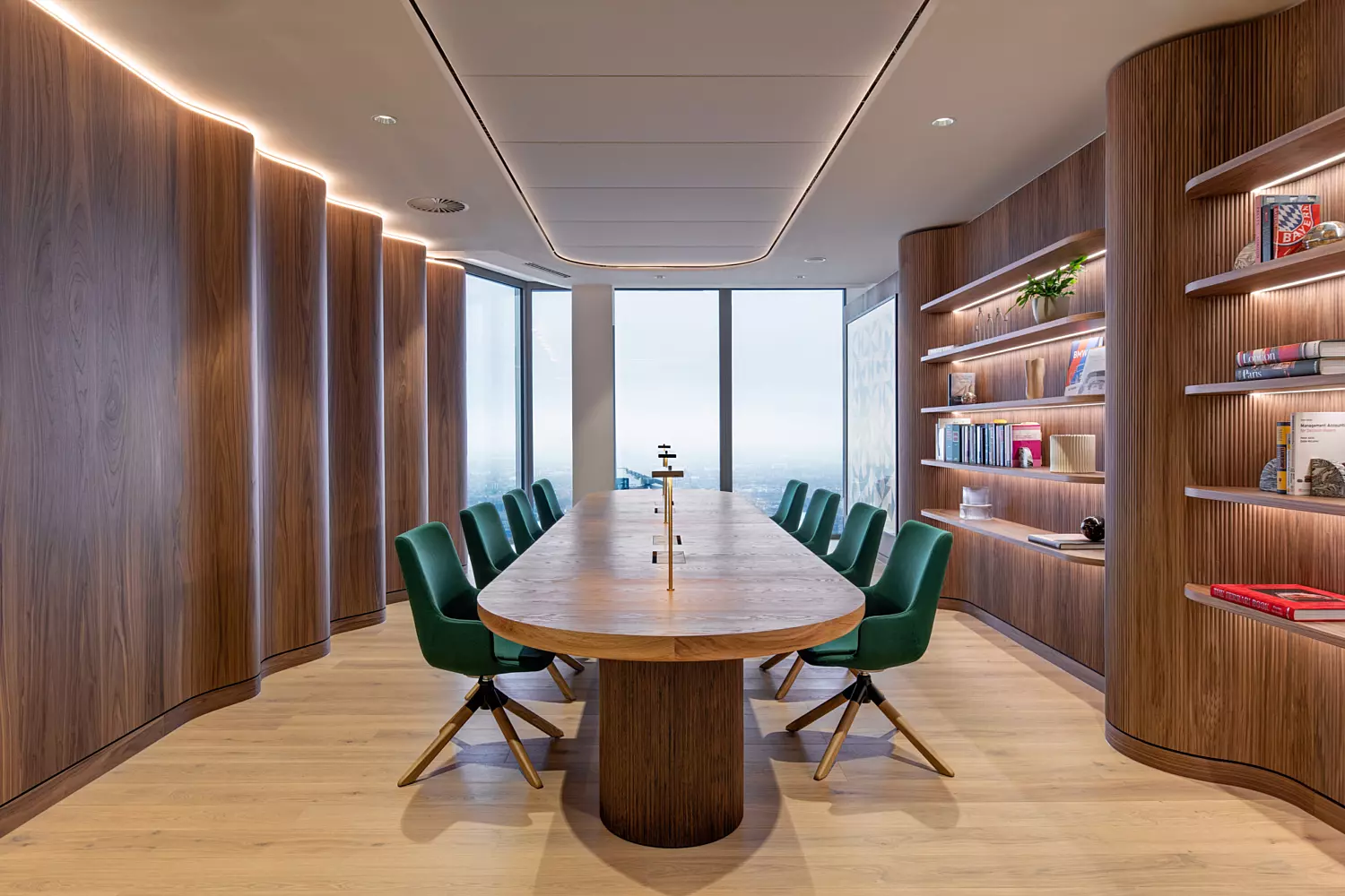A modern conference room featuring a long wooden table surrounded by green upholstered chairs. The walls are clad in wooden panelling, with built-in shelving units displaying books and decorative objects. Large windows at the far end of the room allow natural light to flood the space. The ceiling is fitted with recessed lighting, contributing to the room’s bright and welcoming ambience.