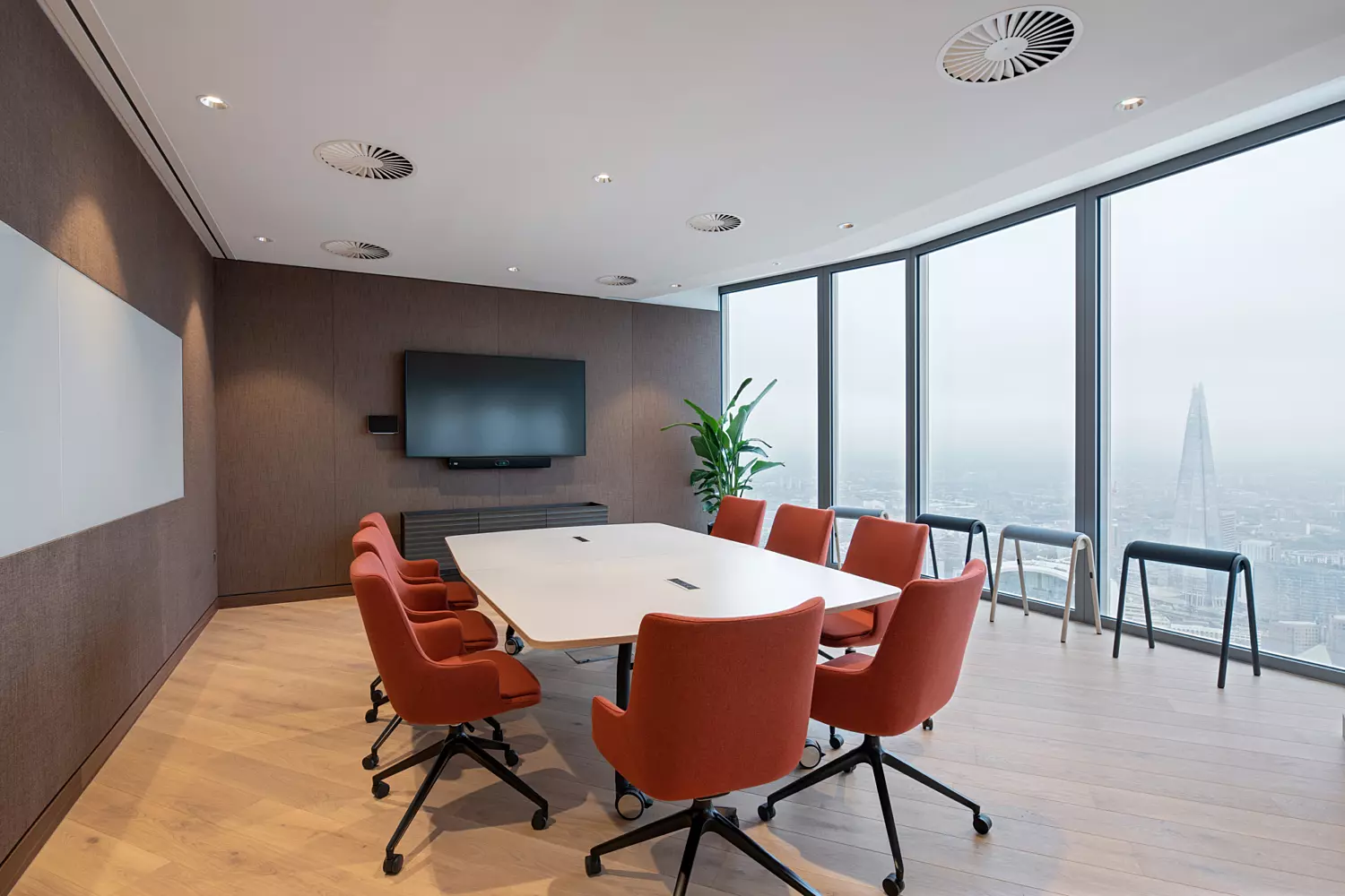 A modern conference room featuring a large white table surrounded by eight orange swivel chairs. Floor-to-ceiling windows provide expansive views of the city skyline, including a prominent skyscraper in the distance. The room includes a wall-mounted flat-screen television and a whiteboard on an adjacent wall. Wooden flooring and ceiling lights contribute to the clean, professional atmosphere. Three high stools are positioned near the windows, and a green plant in the corner adds a touch of nature.