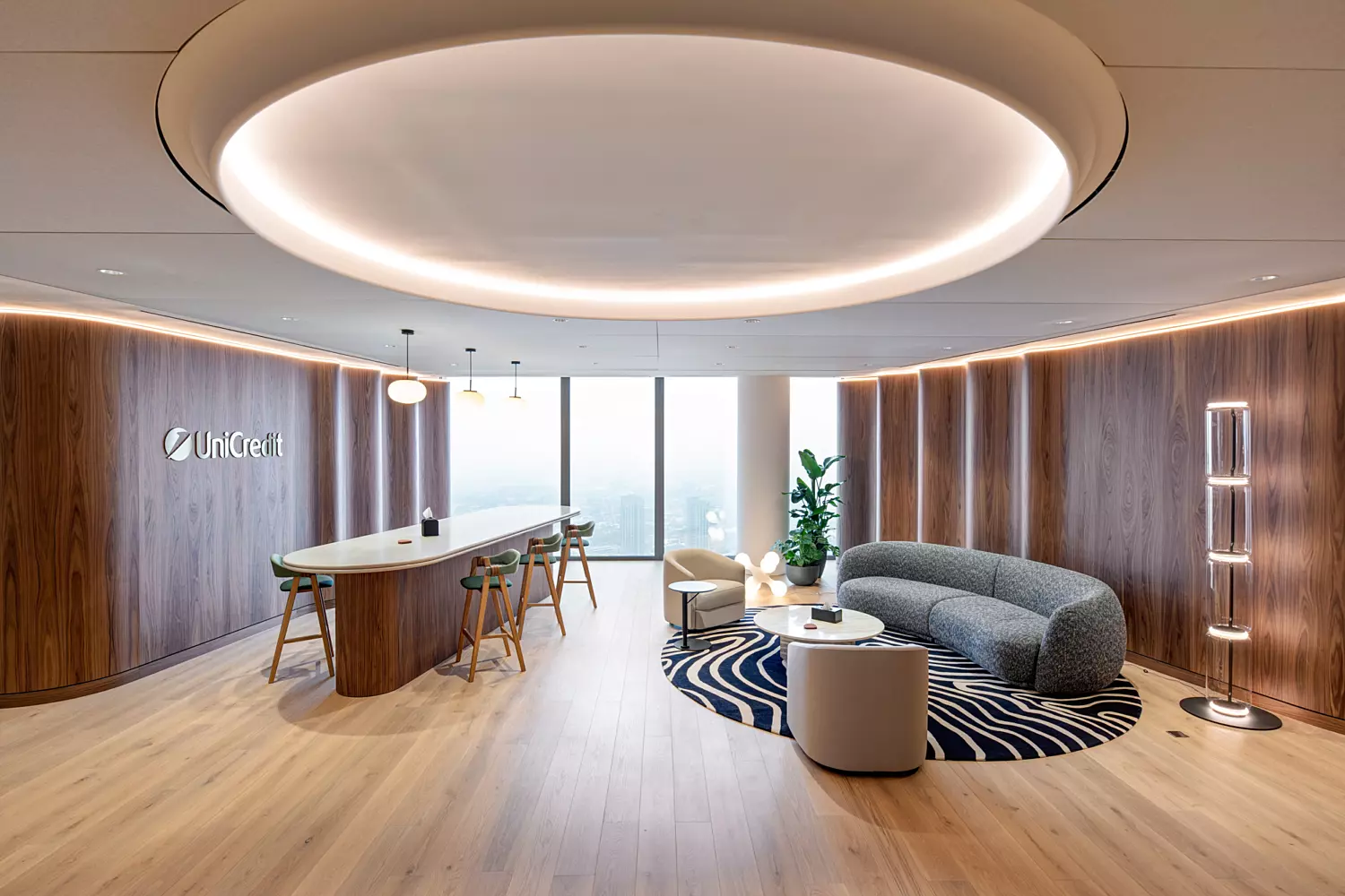A stylish and modern office lounge area featuring a curved grey sofa that forms a semi-enclosed booth-like seating arrangement around a round coffee table placed on a patterned rug. The space includes a sleek oval-shaped bar table with high stools, positioned under a large circular ceiling light fixture. Wooden flooring and wall paneling add warmth to the environment, while a tall indoor plant in the corner and large windows offering natural light enhance the inviting atmosphere. The word "UniCredit" is displayed on the wall to the left, contributing to the branded interior design.