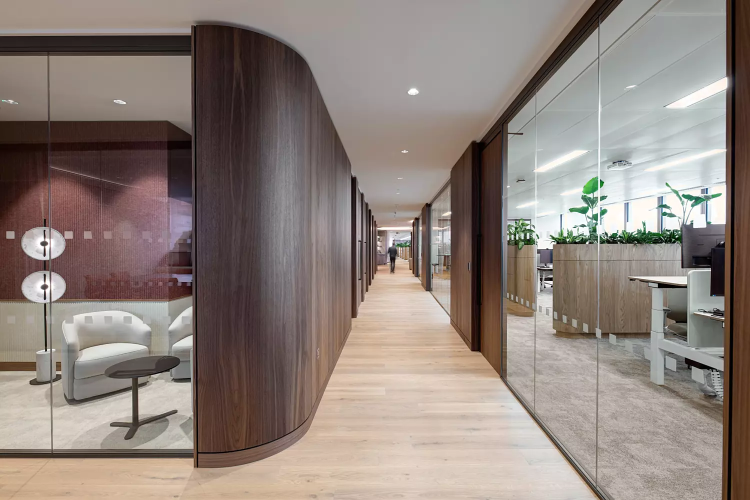 A contemporary office corridor with wooden flooring and wall panelling. On the left, glass-walled meeting rooms are visible, one containing white chairs and a small table. To the right, glass partitions separate the hallway from an open-plan office area furnished with desks and decorated with green plants. The space is brightly lit with ceiling lights, and a person is seen walking at the far end of the corridor.