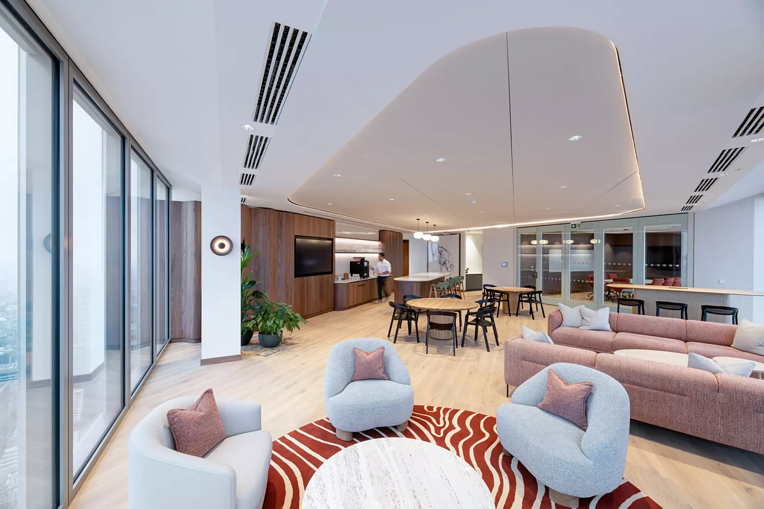 A modern, open-plan living space with large windows along the left side, allowing natural light to fill the room. The foreground features a seating area with three light grey armchairs arranged around a round white marble coffee table, set atop a red and white patterned rug. In the background, a dining area with several tables and chairs leads into a kitchen or bar area fitted with wooden cabinetry. The ceiling includes recessed lighting and a distinctive architectural design, while potted plants near the windows add a touch of greenery.