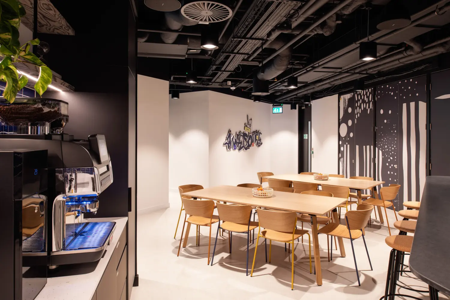 Modern office café and breakout area featuring a self-service coffee station, light wood dining tables and contemporary chairs. The space includes banquette seating, feature wall graphics and decorative patterned partitions, with exposed black ceilings and pendant lighting creating a relaxed setting for informal meetings, refreshments and collaboration.