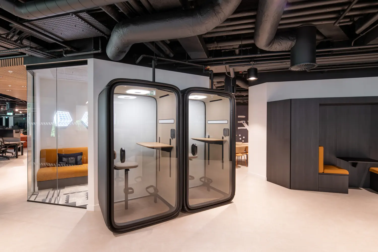 Modern office collaboration space featuring two enclosed acoustic phone booths with standing-height work surfaces and stools. The area includes a glass-fronted meeting room, built-in booth seating with mustard-coloured upholstery and an open-plan workspace beyond. Exposed black services, contemporary lighting and clean, minimalist finishes create a modern and flexible working environment.