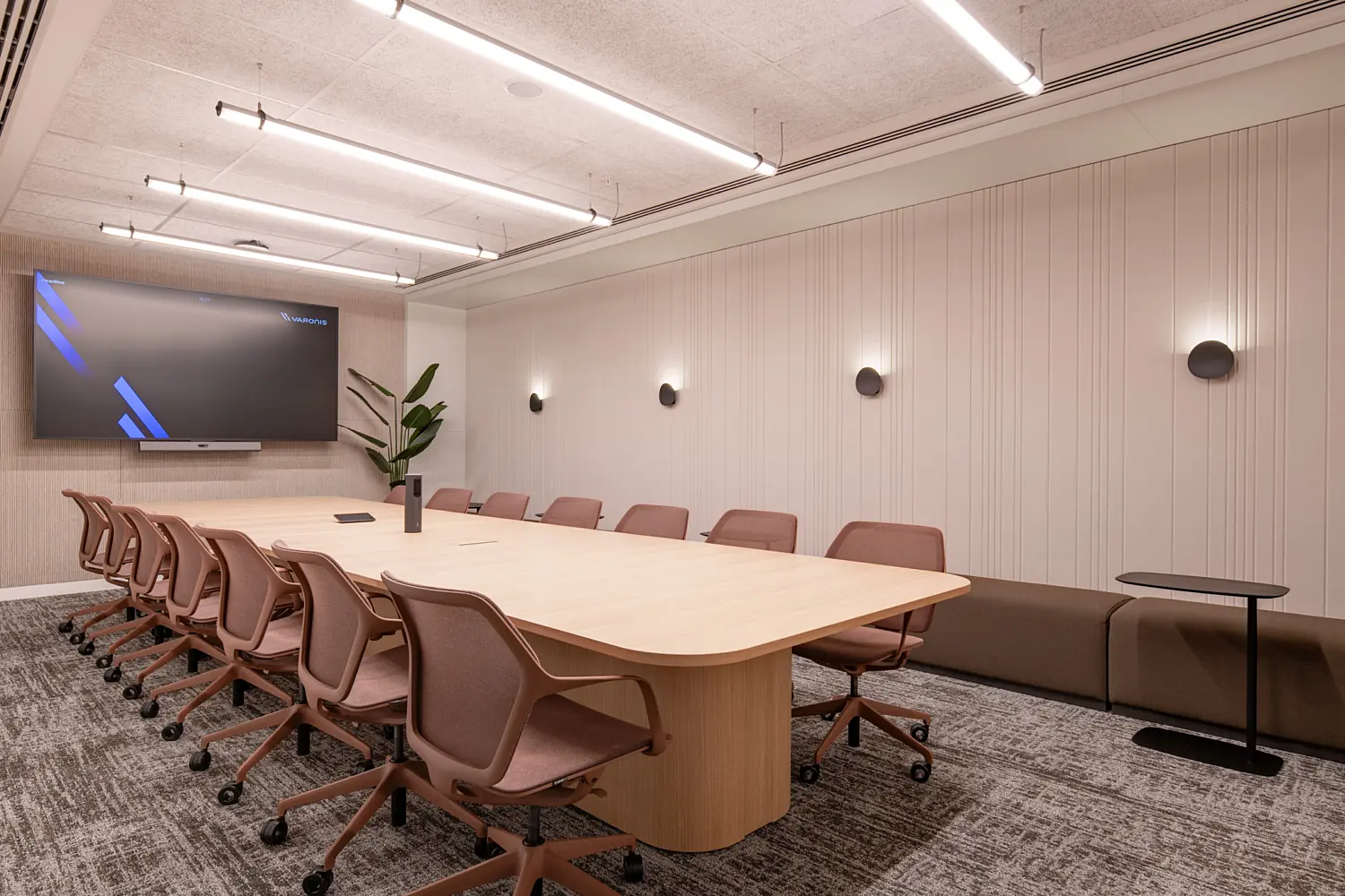 Contemporary boardroom featuring a large light oak conference table surrounded by ergonomic meeting chairs in a muted terracotta tone. A wall-mounted display screen sits at one end of the room, while acoustic wall panelling, integrated lighting and neutral finishes create a clean, professional meeting environment. A built-in bench seat and planting add warmth and comfort to the space.
