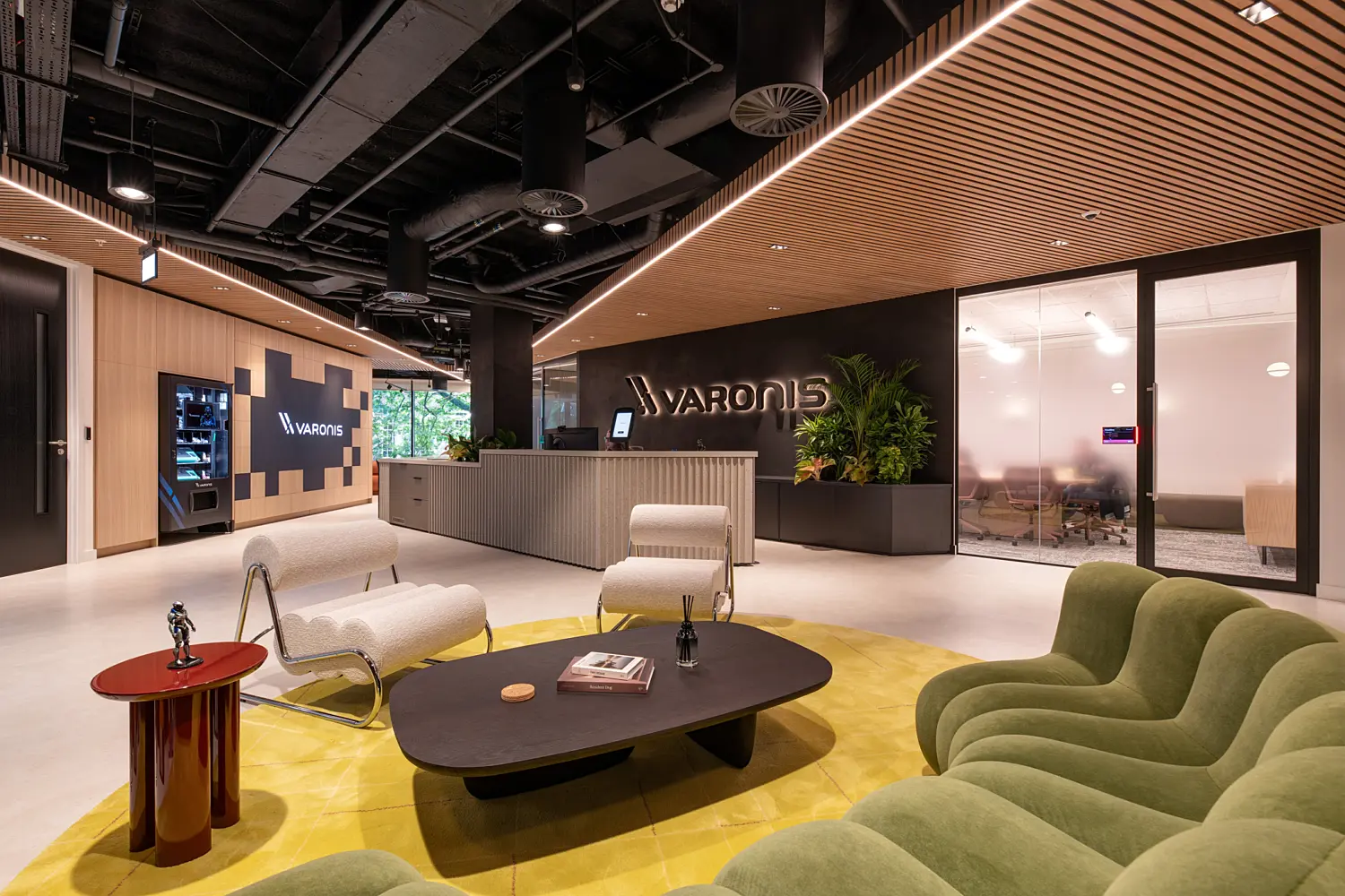 Modern office reception area featuring a central welcome desk with illuminated Varonis branding, surrounded by contemporary lounge seating. The space includes a curved green sofa, upholstered chairs, circular coffee tables and indoor planting, with warm timber slatted ceilings, black exposed services and glass-fronted meeting rooms creating a sleek, high-end workplace environment.
