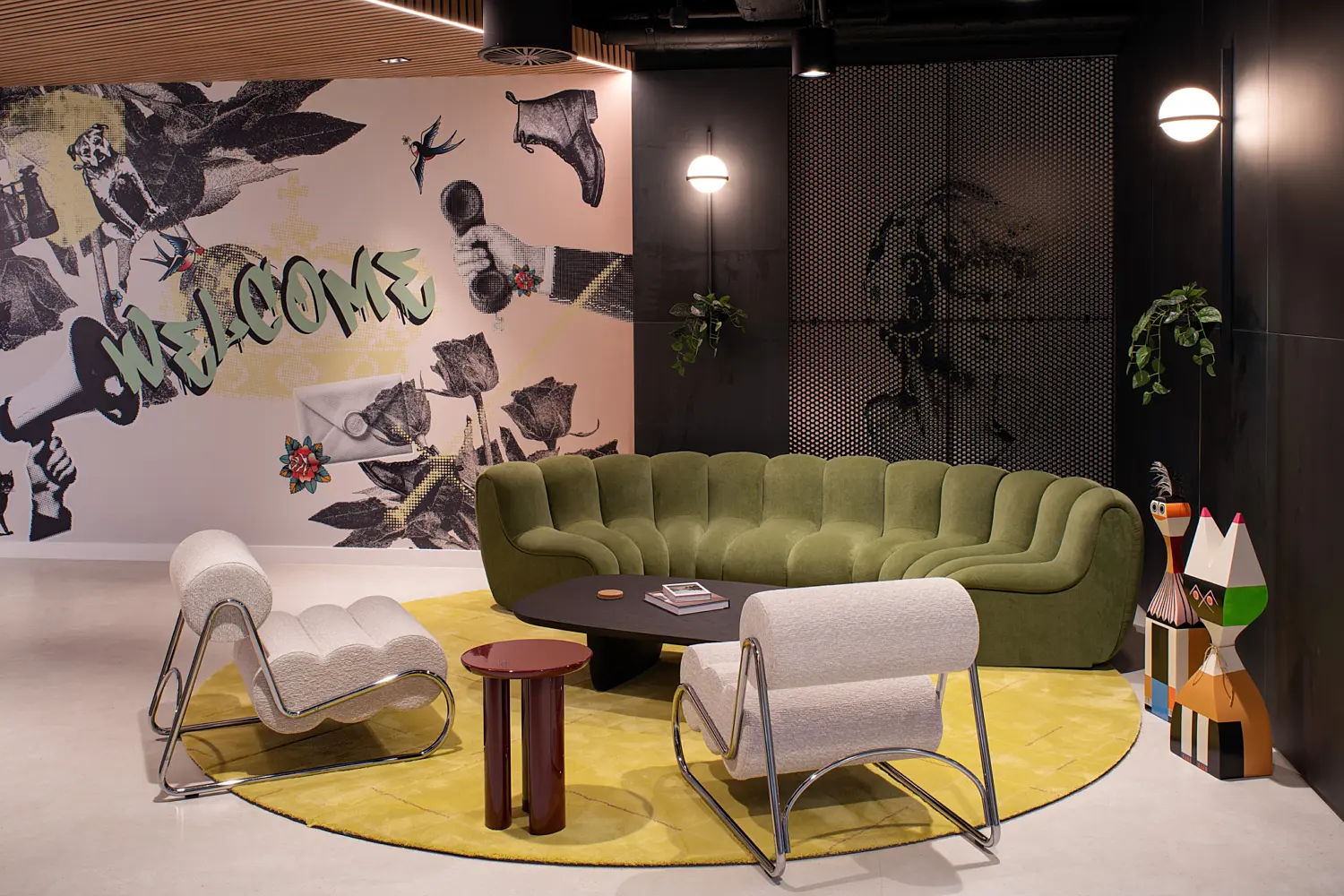 Contemporary office lounge area featuring a sculptural green curved sofa, two white accent chairs and a black coffee table arranged on a circular yellow rug. A large feature mural with mixed-media artwork and the word "WELCOME" forms the backdrop, while dark walls, timber slatted ceiling details, decorative lighting, planting and colourful statement pieces create a bold and inviting reception space.