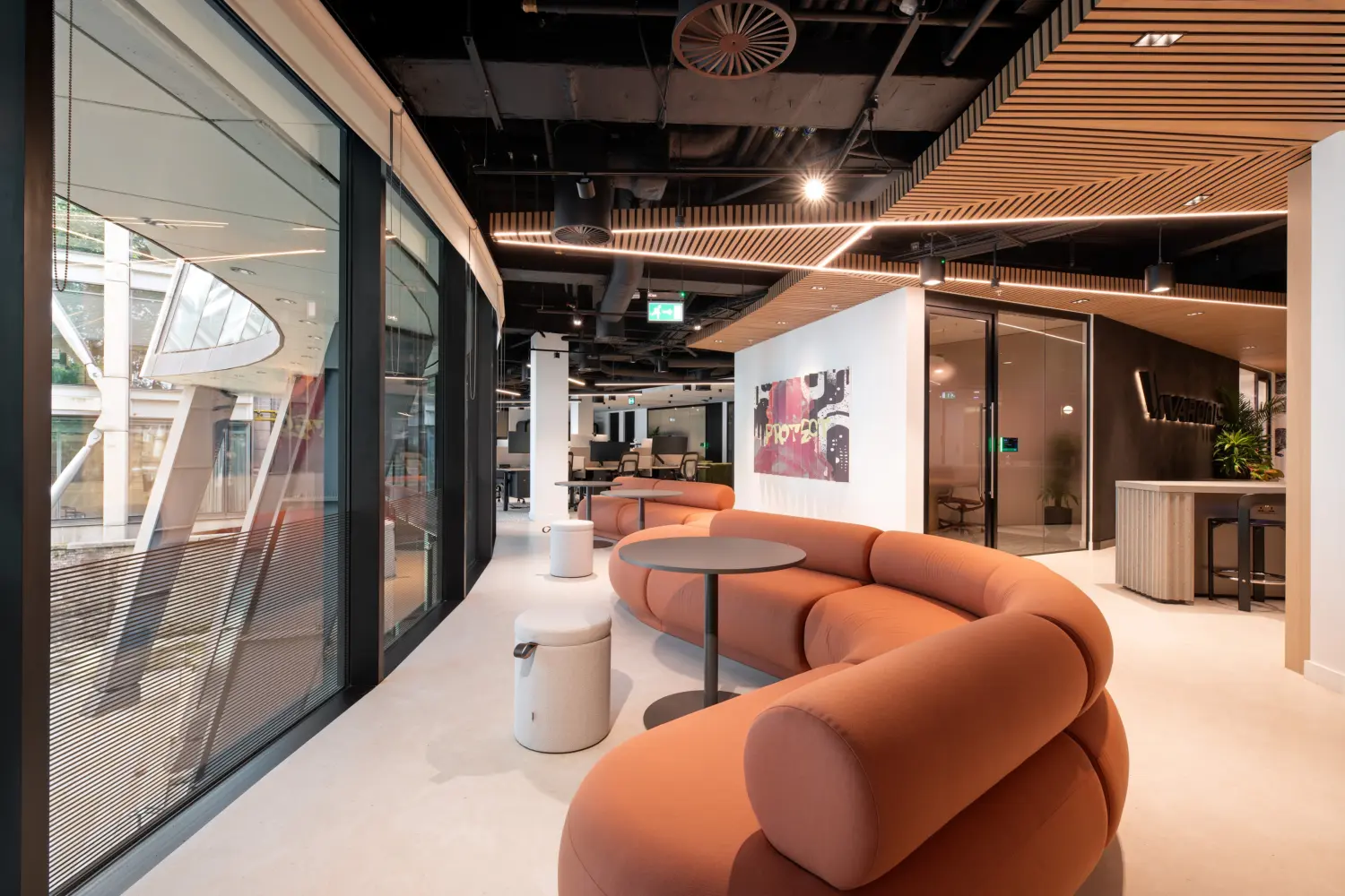 Modern office lounge area featuring a curved terracotta-coloured sofa and round tables positioned alongside floor-to-ceiling glazing. The space leads into an open-plan workplace with meeting rooms, collaborative seating and a reception area displaying Varonis branding. Timber slatted ceiling features, exposed black services and warm lighting create a contemporary, high-specification office environment.