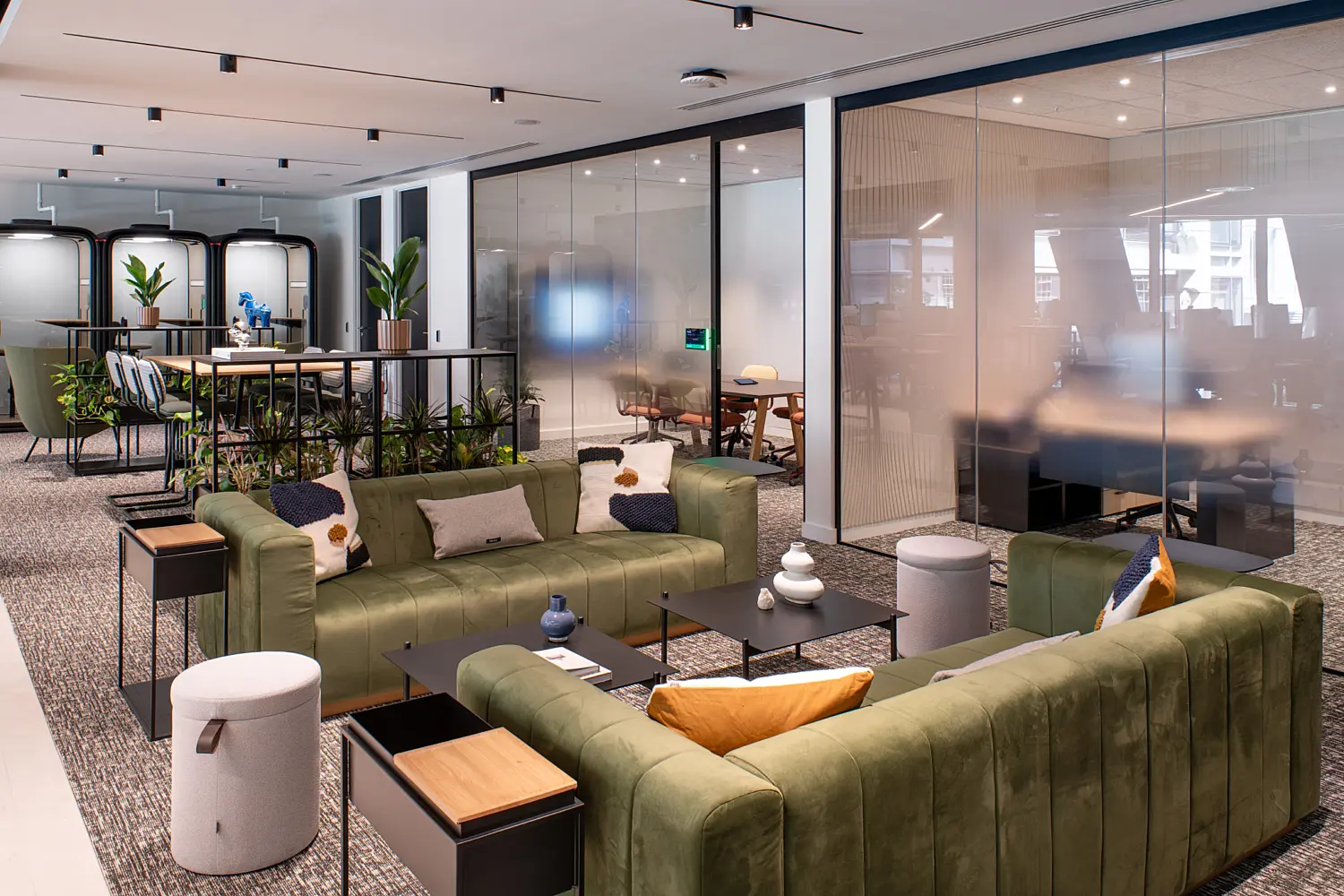 Contemporary office breakout and collaboration area featuring two green sofas arranged around black coffee tables, with adjacent informal seating and meeting spaces. Floor-to-ceiling glass partition walls enclose meeting rooms, while planters, soft furnishings and modern furniture create a comfortable workspace with a neutral colour palette and abundant natural light.