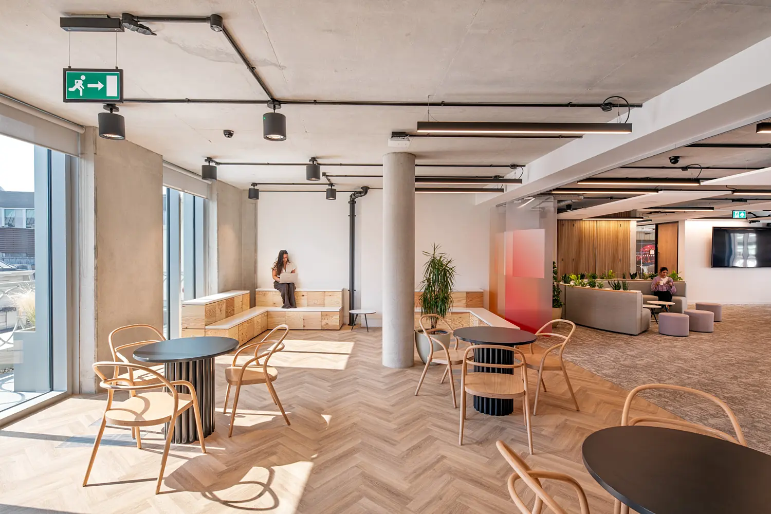 Bright open-plan office with floor-to-ceiling windows, herringbone timber flooring and exposed concrete ceilings. Round black tables with timber chairs provide informal seating, while tiered wooden bench seating runs along the windows. Two people are seated in separate areas. Planting, a grey sofa, small stools and a timber slatted wall can be seen further into the workspace.