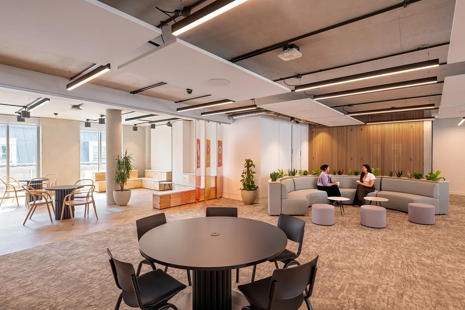 Open plan office with a round meeting table and chairs in the foreground. A curved grey sofa and small stools form an informal seating area, where two people are seated facing each other. Large windows, exposed concrete columns, indoor planting and a timber slatted wall add warmth to the space, while linear ceiling lights run across the exposed concrete ceiling.