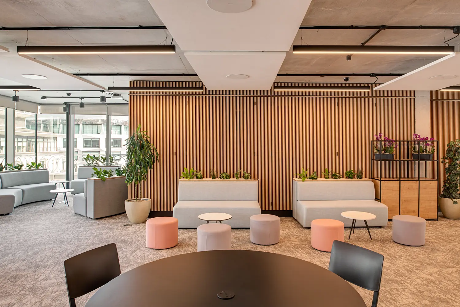 Informal office breakout area with grey sofas and pastel stools arranged around small round tables. Integrated planters and potted plants add greenery, set against a timber slatted wall. A large round black table sits in the foreground, while floor-to-ceiling windows bring natural light into the space. Exposed concrete ceilings and linear lighting complete the interior.