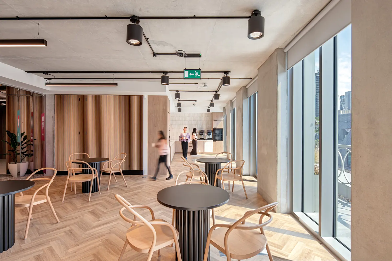 Bright office breakout area with round black tables and light timber chairs beside floor-to-ceiling windows. Herringbone timber flooring, exposed concrete columns and ceilings give the space an industrial finish, with black ceiling-mounted lighting overhead. A timber slatted wall and indoor plants sit to the left, while three people are visible moving through and standing in the background near a kitchenette.