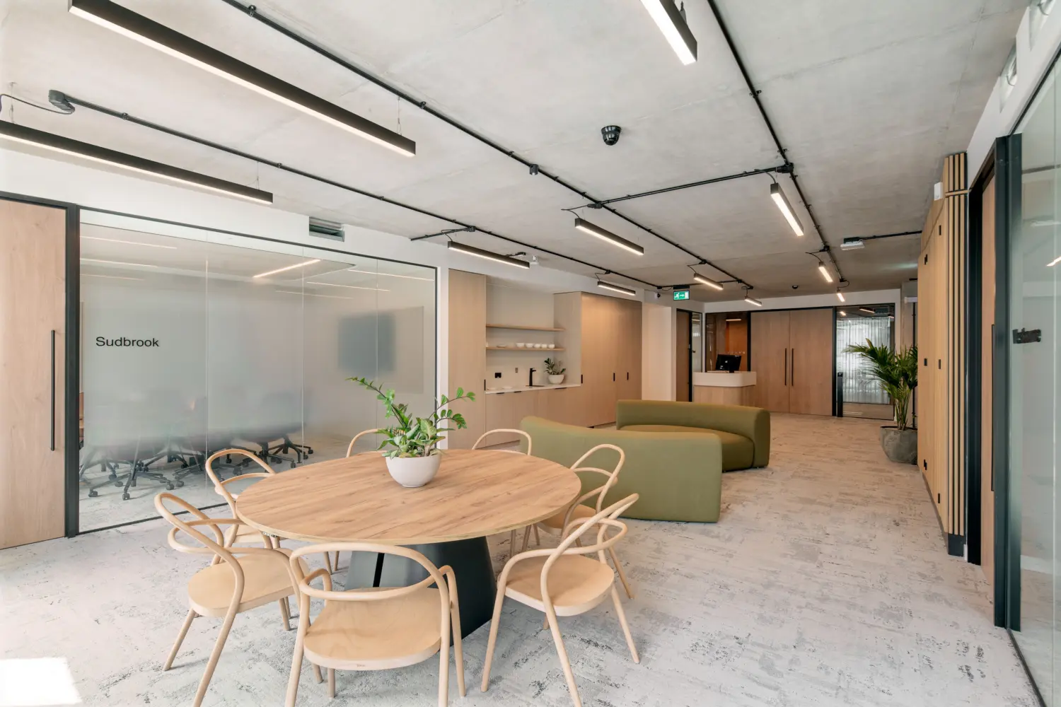 Open office breakout space with a large round timber table and light wooden chairs in the foreground. Green upholstered seating sits beyond, alongside pale timber storage, shelving and a reception desk. A glass-fronted meeting room is on the left. Exposed concrete ceilings, linear lights, timber finishes, indoor planting and neutral carpet complete the space.