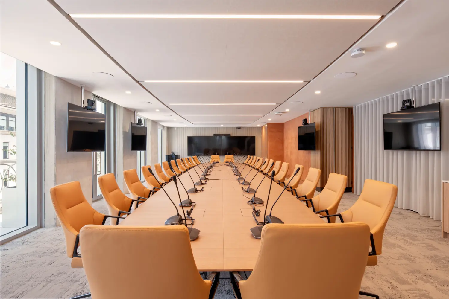 Large boardroom with a long U-shaped timber conference table surrounded by orange chairs. Individual microphones are positioned around the table, with several wall-mounted screens and cameras for video conferencing. Floor-to-ceiling windows run along one side, while timber panelling and white curtains line the opposite walls. Linear ceiling lights illuminate the room.