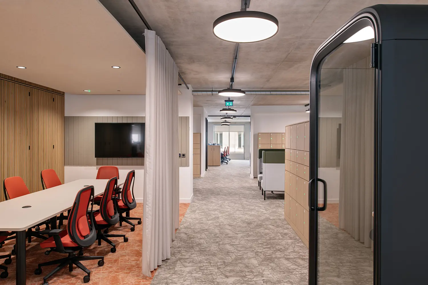 Modern office corridor with an exposed concrete ceiling and circular pendant lights. A meeting area with a long table, red office chairs, wall-mounted screen and timber slatted wall sits to the left, separated from the corridor by a curtain. Lockers and work booths line the right side, with a dark framed glass meeting pod in the foreground.