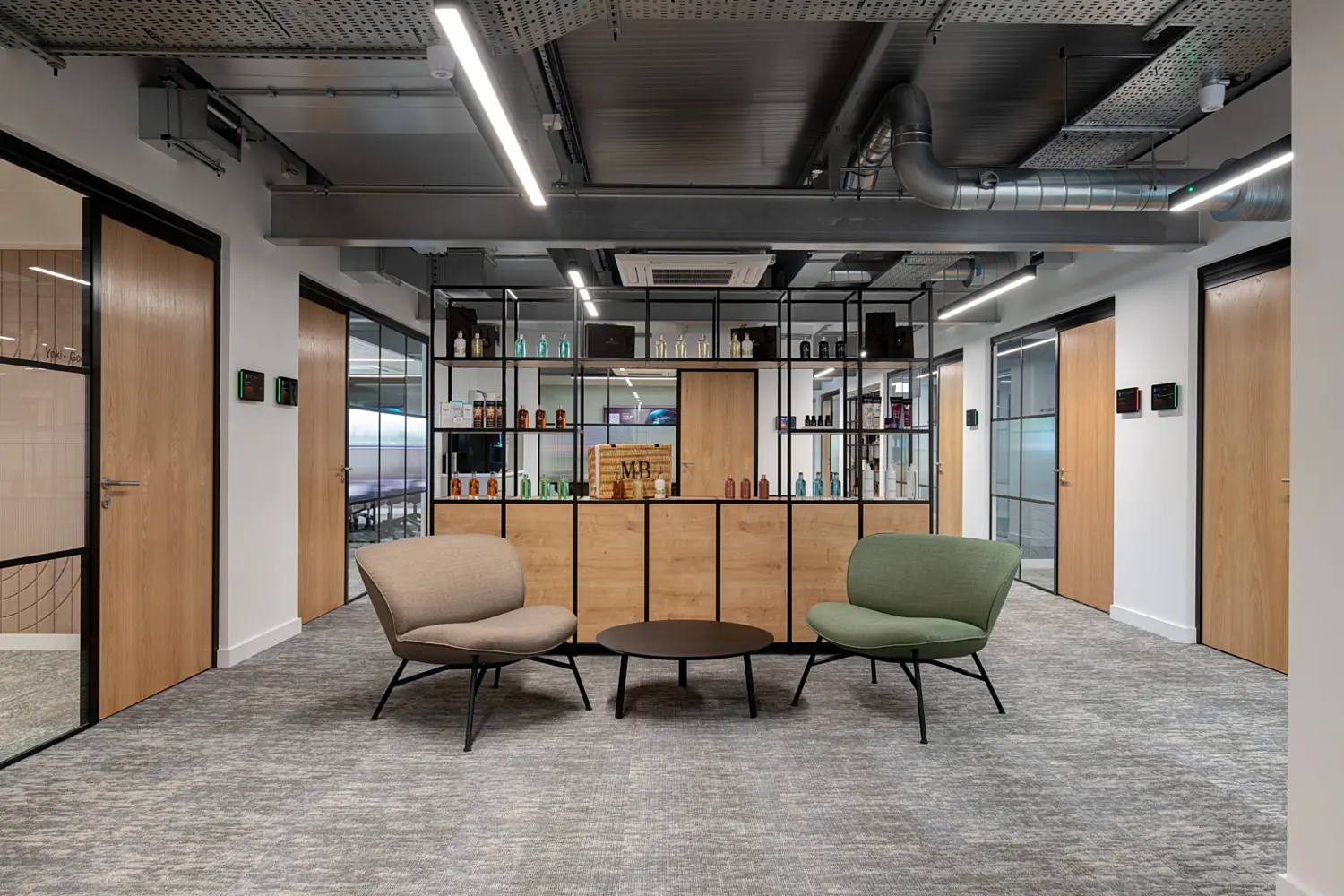 A contemporary office lounge area with two low armchairs, one in beige and one in green, arranged around a small circular coffee table on a grey patterned carpet. Behind them, a floor to ceiling shelving unit with black framing and wood panels displays assorted bottles and decorative items. Four timber doors line the surrounding white walls, and the ceiling features exposed ductwork, cable trays and linear LED lighting.