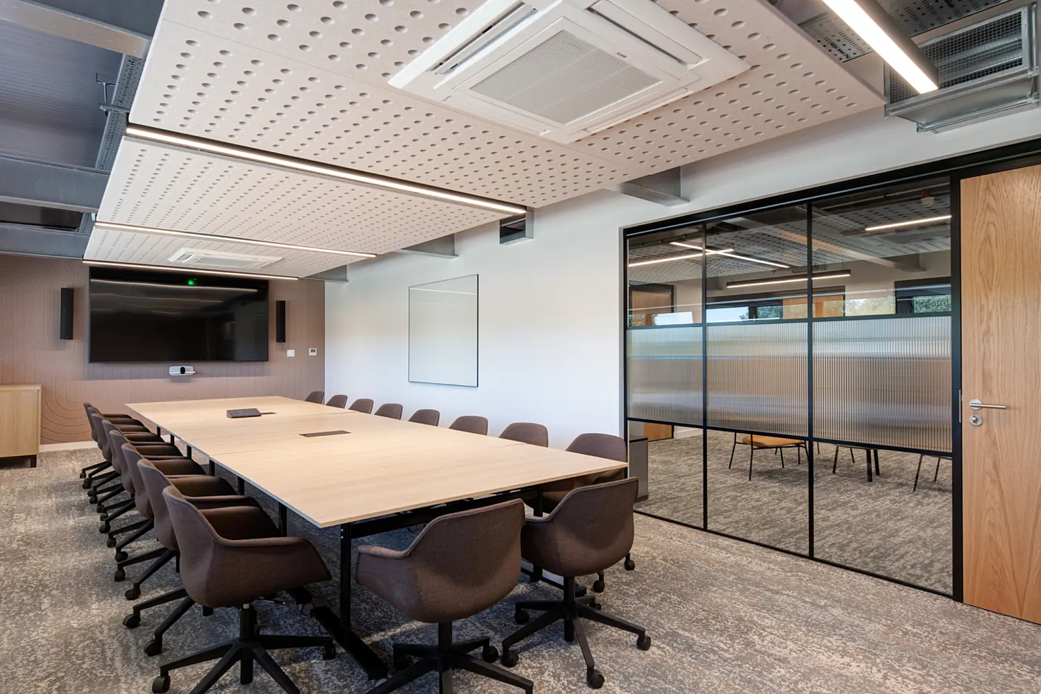 A modern meeting room with a large rectangular light wood table surrounded by swivel chairs upholstered in dark brown fabric. A wall mounted screen and video conferencing unit sit at the far end of the room above a wood credenza. The space has a grey patterned carpet and a ceiling with exposed services and perforated acoustic panels. To the right, a glass partition with black framing and fluted panels separates the room from an adjacent workspace, with a timber door set into the partition.Provide your feedback on BizChat