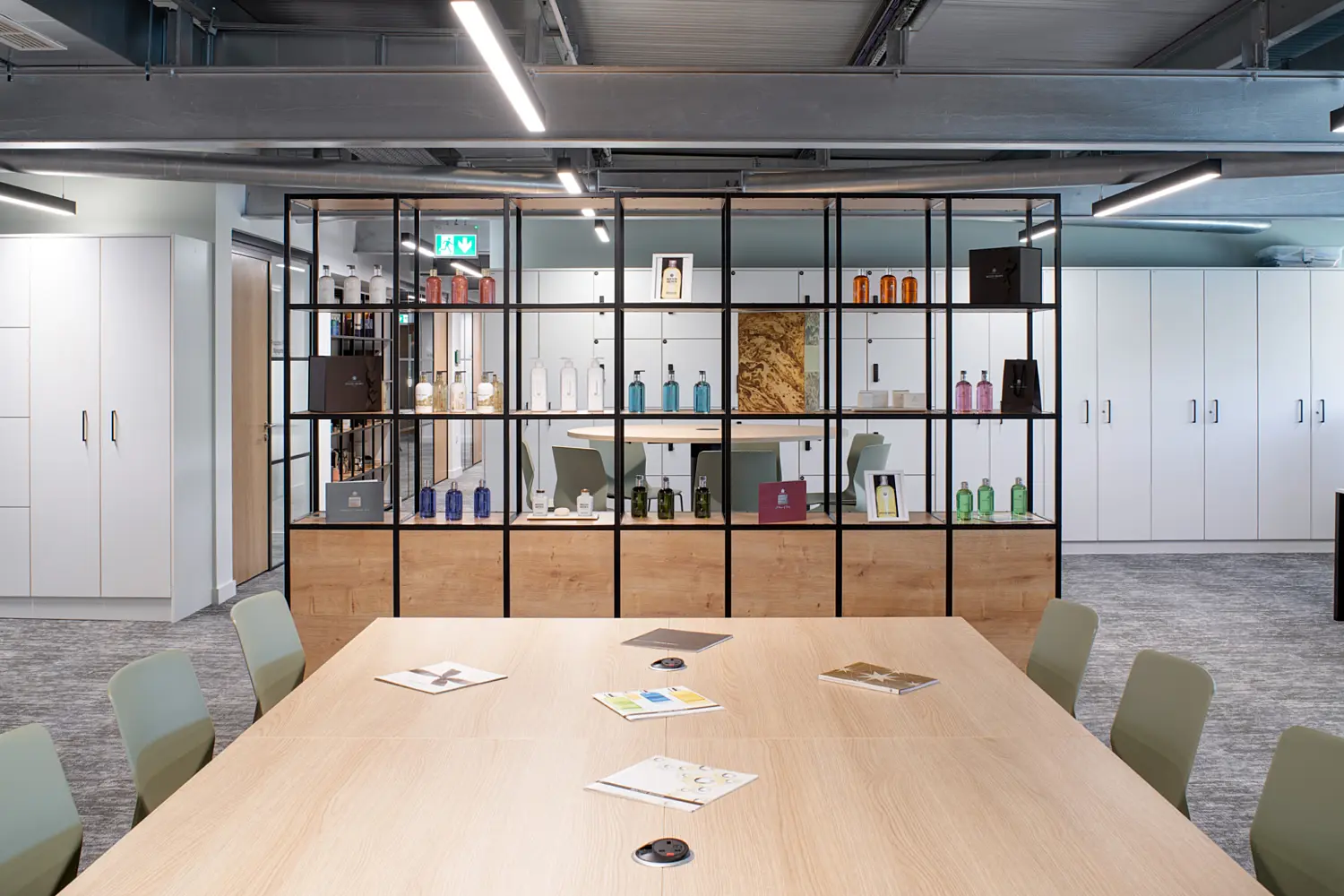 A meeting room with a large light wood table surrounded by green chairs, with notebooks and documents placed on the surface. Behind the table, a floor to ceiling shelving unit with a black metal frame and wood panels displays assorted bottles, packaging and framed items. White storage cabinets line the walls on either side. The ceiling has exposed services and linear LED lighting, creating a bright and open workspace.