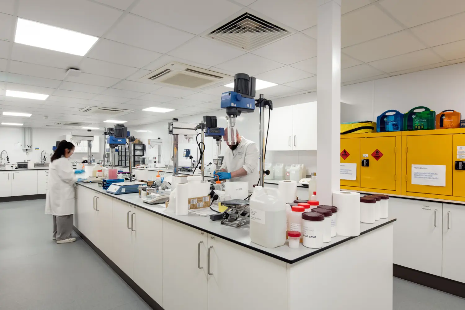 A bright laboratory with white walls, ceiling panels and long central workbenches. Containers, instruments and mixing equipment are arranged across the surfaces. Two people in lab coats and gloves are working at separate stations, one using a bench mixer and the other preparing materials. Safety cabinets in bright yellow stand against the right wall, and various pieces of technical equipment and storage line the background.